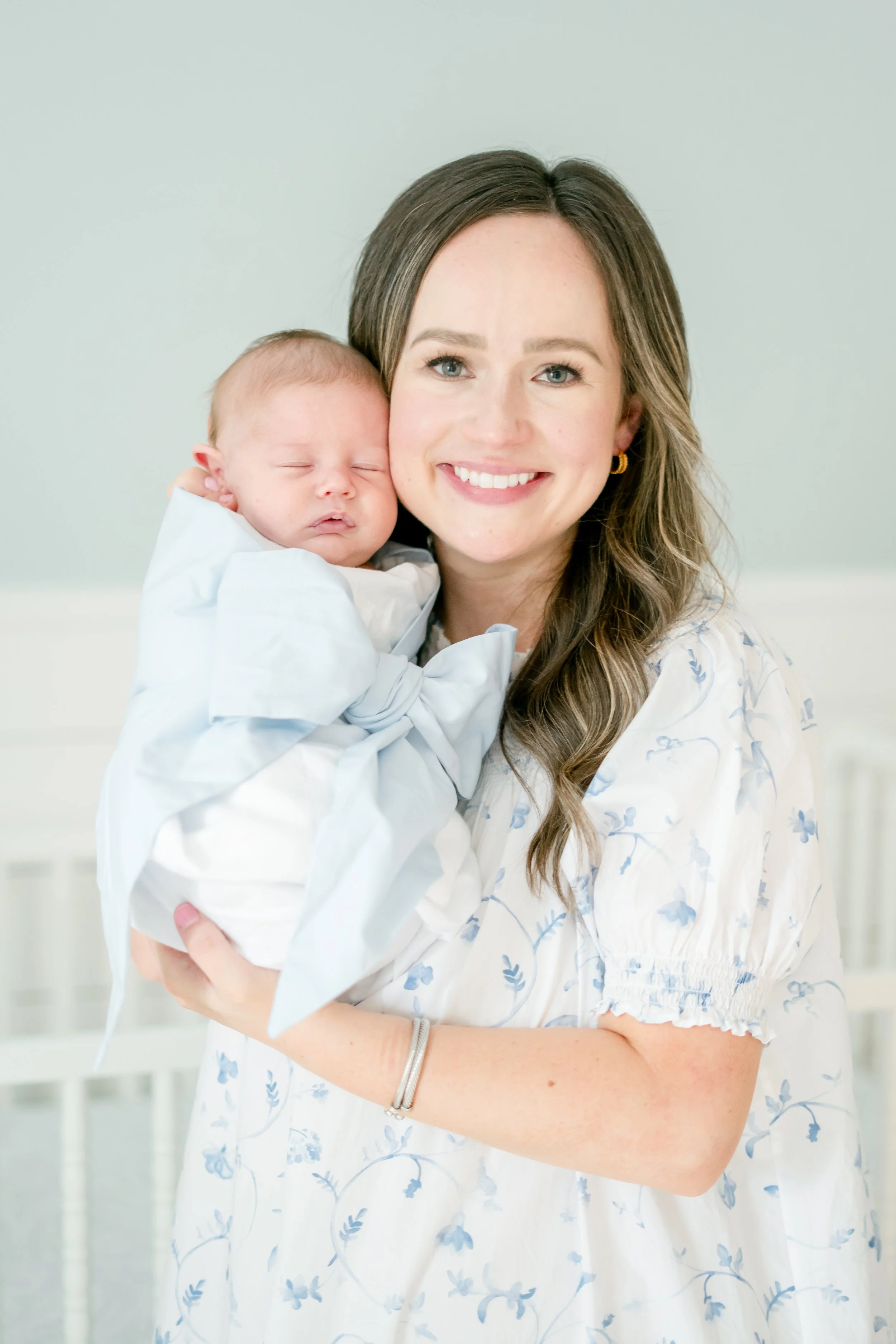 A woman in a white dress with blue floral patterns holding a sleeping baby dressed in white, both smiling, in a light-colored room.