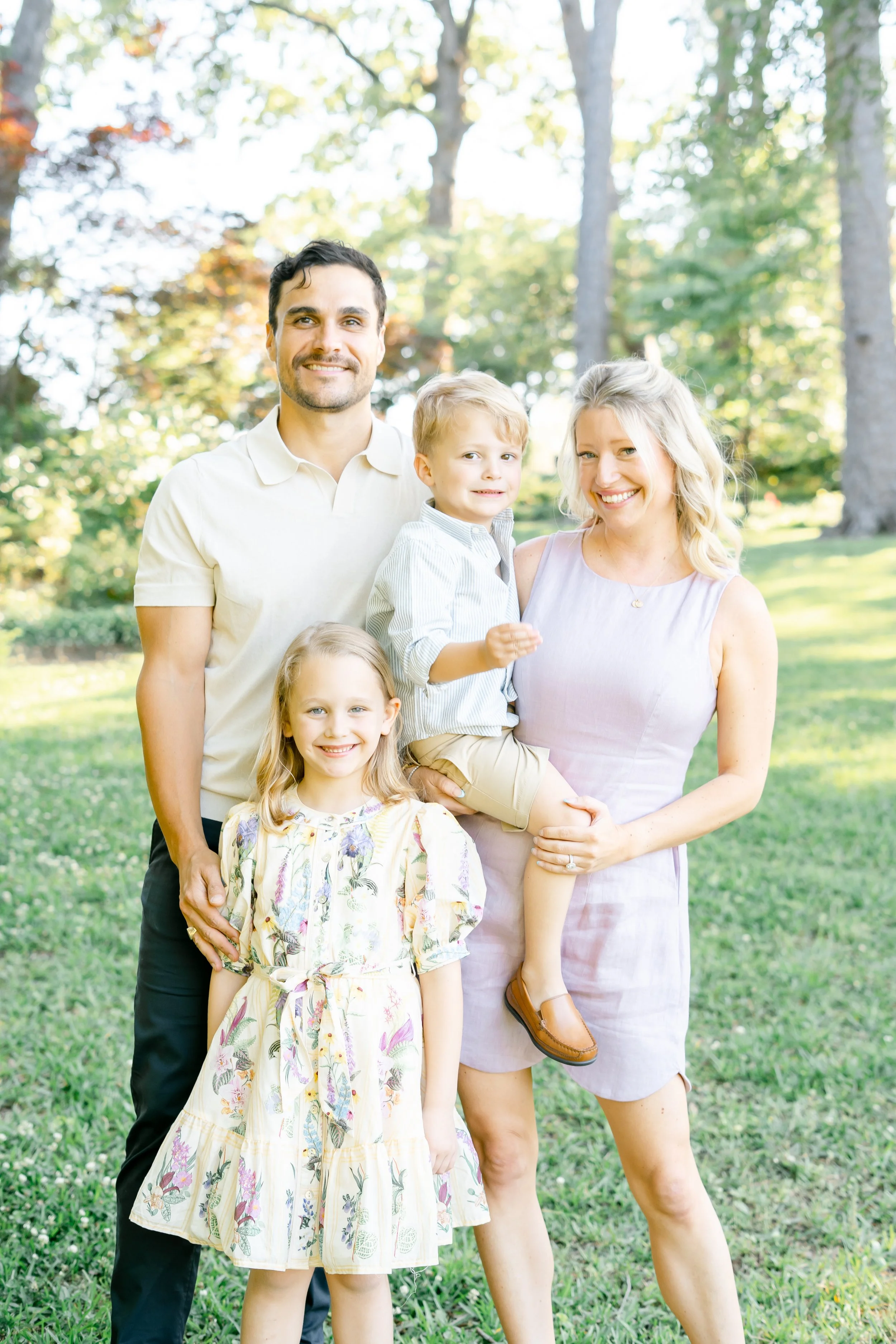 A family of five standing outside in a park with trees in the background. The mother is holding a young boy on her hip, and a young girl is standing in front of her. A man is standing to the left, and partially visible at the bottom is a small child.