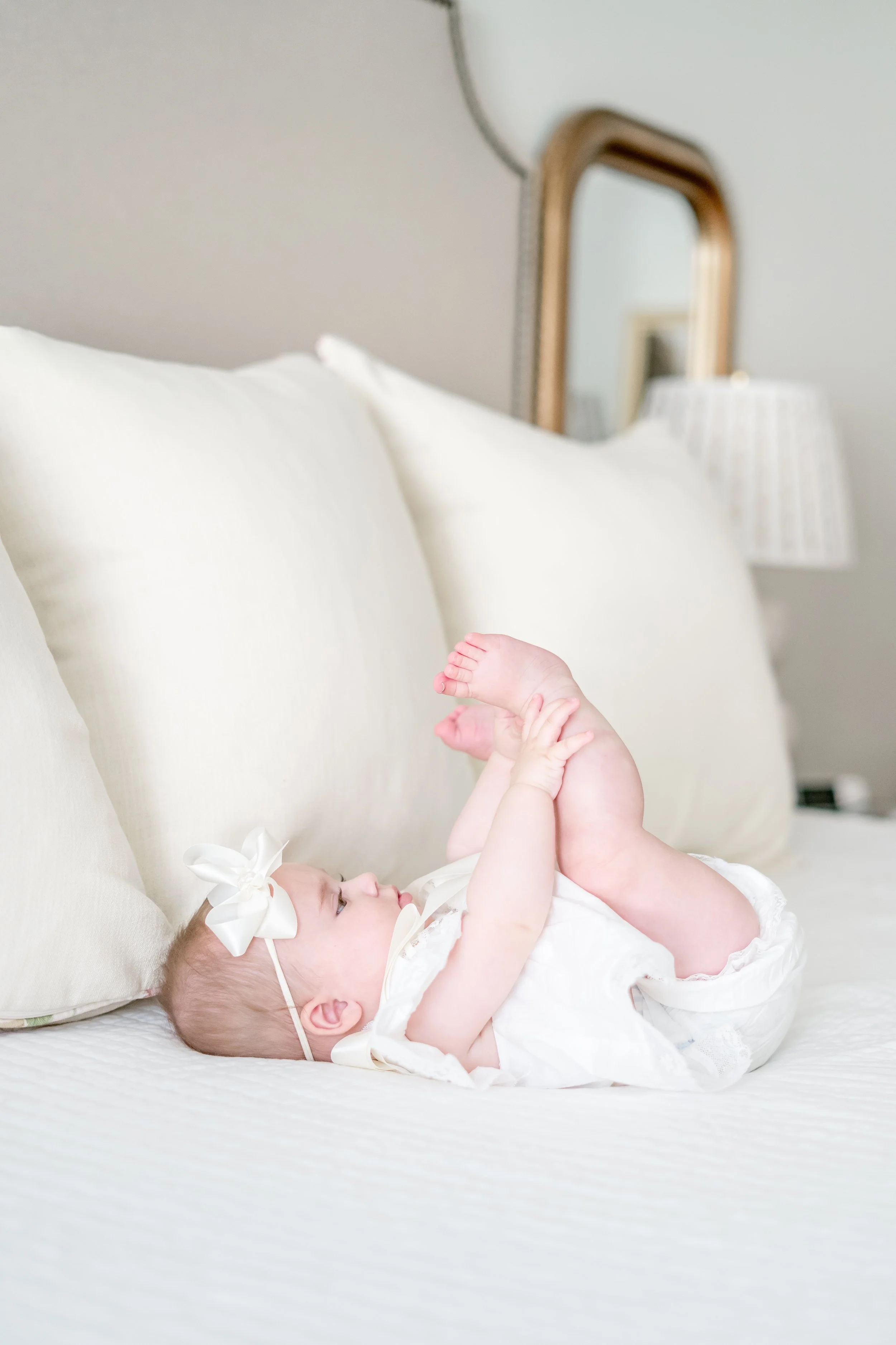 A baby lying on a bed with pillows, playing by holding her feet, wearing a white dress and a large white bow headband.