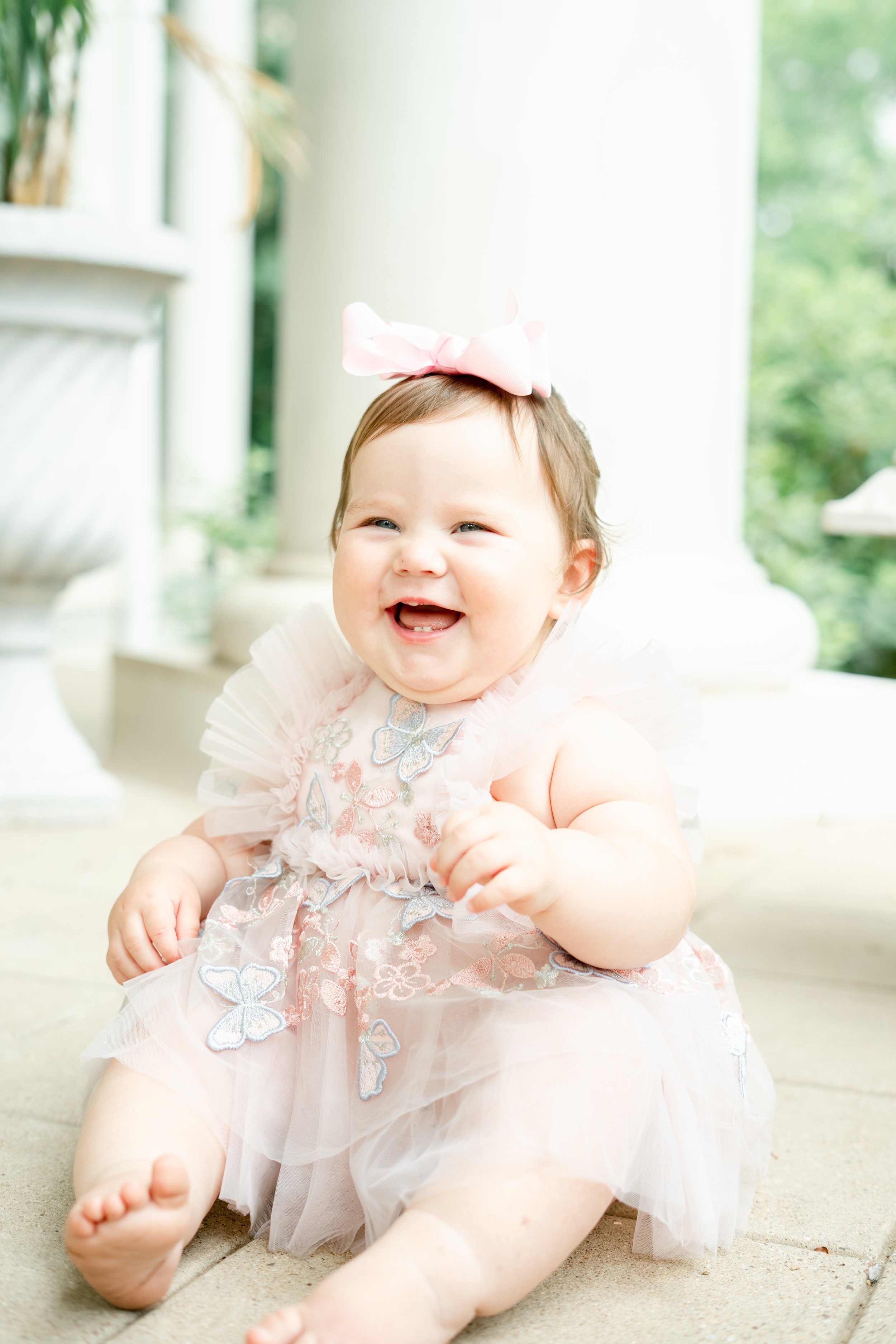 A smiling baby girl wearing a pink dress with embroidered butterflies and a pink bow headband, sitting on a patio with greenery outside and a white column behind her.