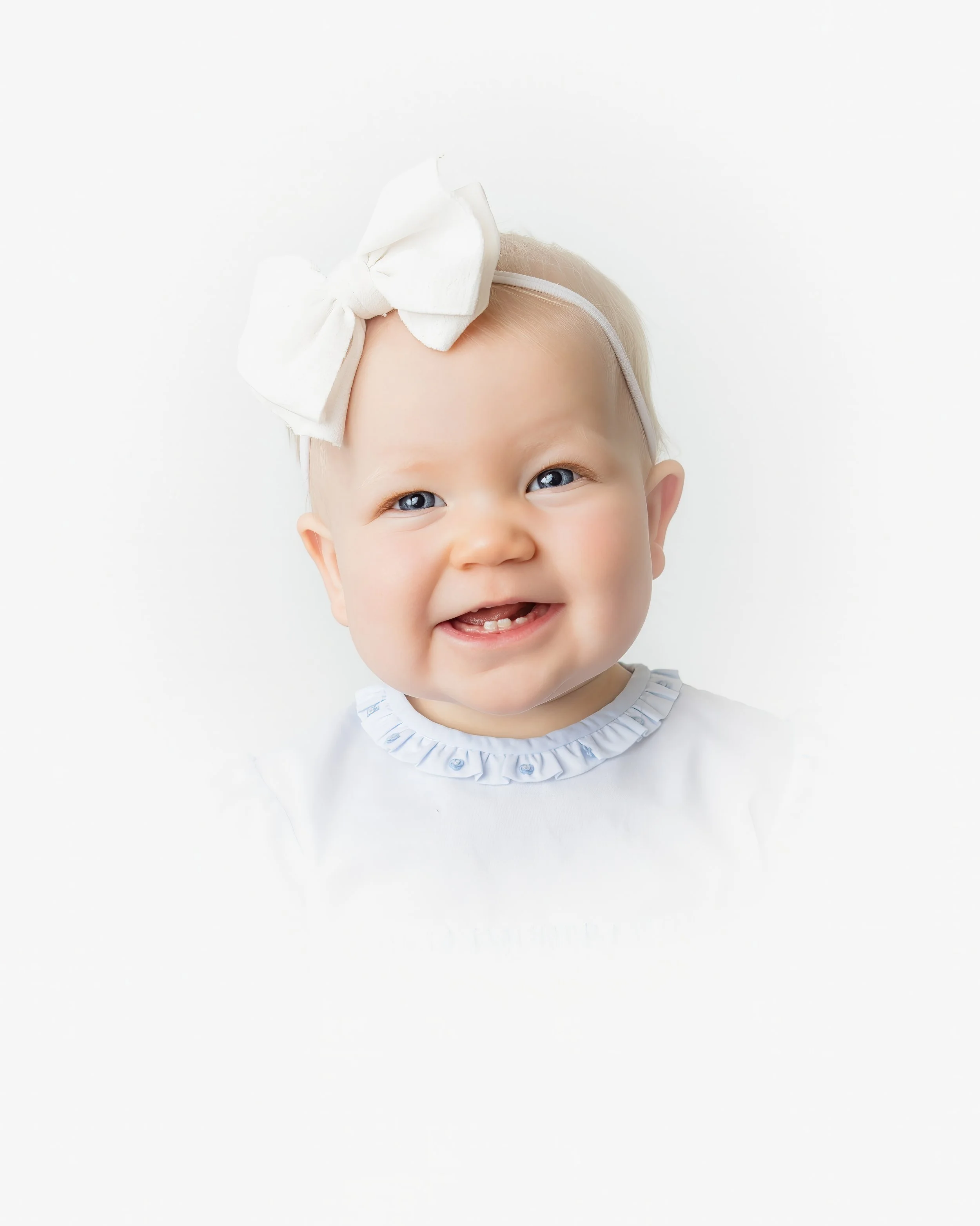 Close-up of a smiling baby girl with blue eyes and light hair, wearing a white headband with a large bow and a white outfit with ruffled collar, against a plain white background.