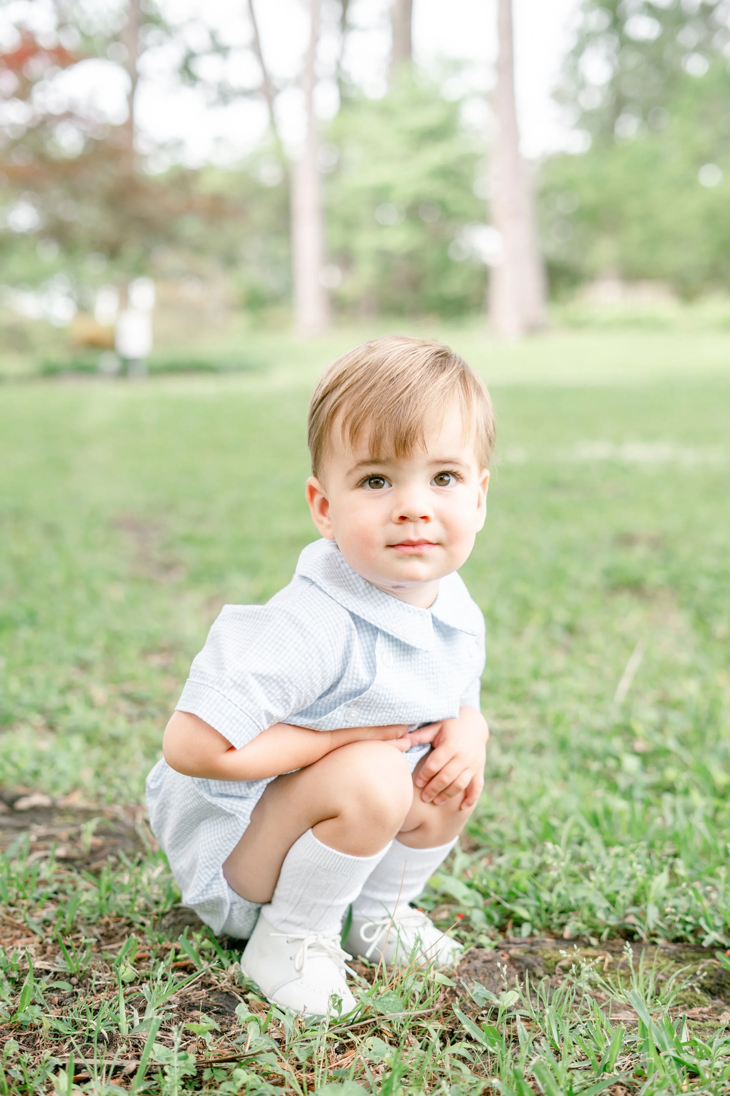 A young boy with blond hair and blue eyes, wearing a light plaid shirt, squatting outdoors on grass, surrounded by trees.