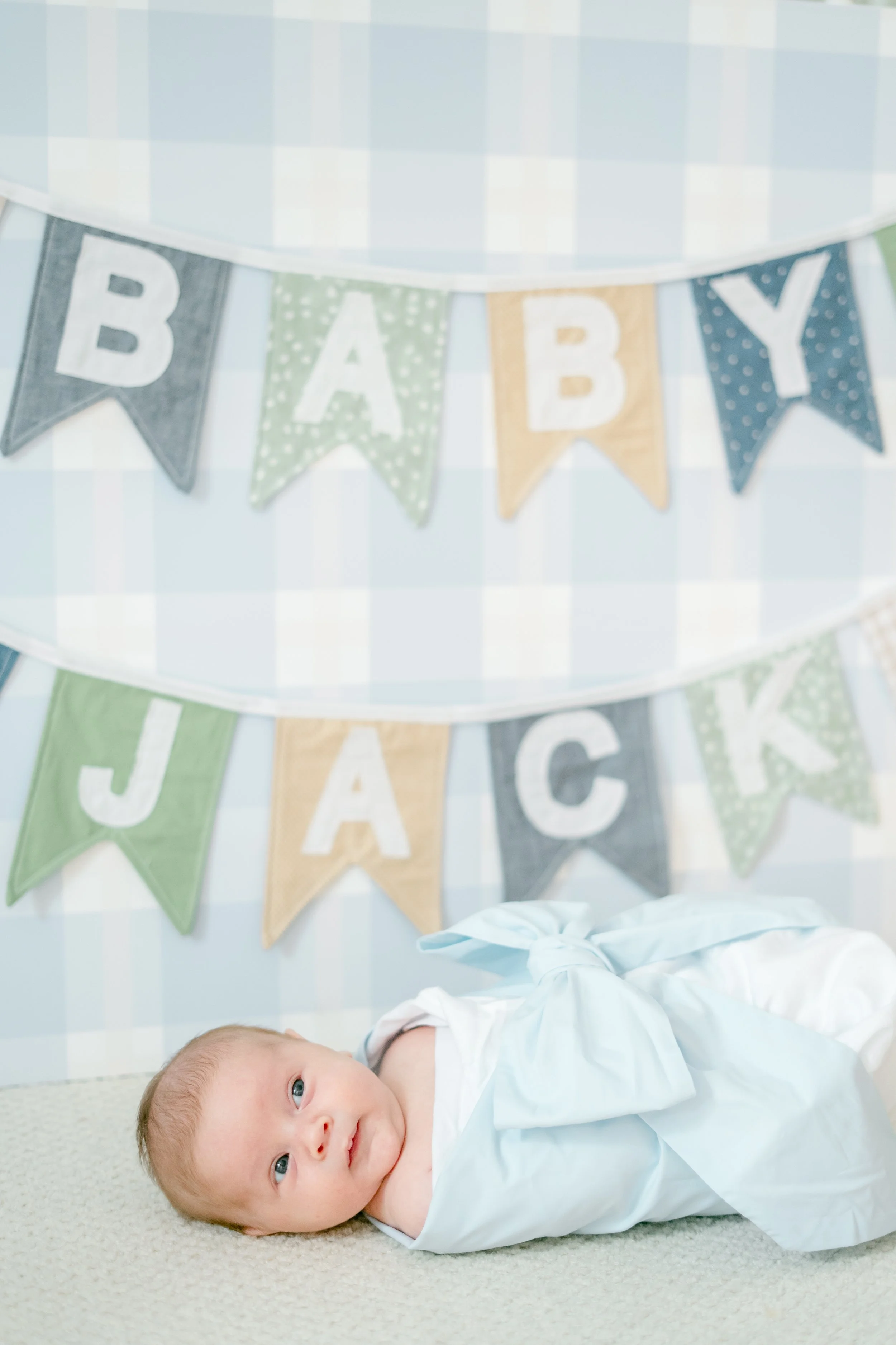A baby with light skin and blue eyes lying on a beige carpet, wearing a white long-sleeve gown, with a backdrop of pastel-colored 'BABY' banners hanging on a wall.