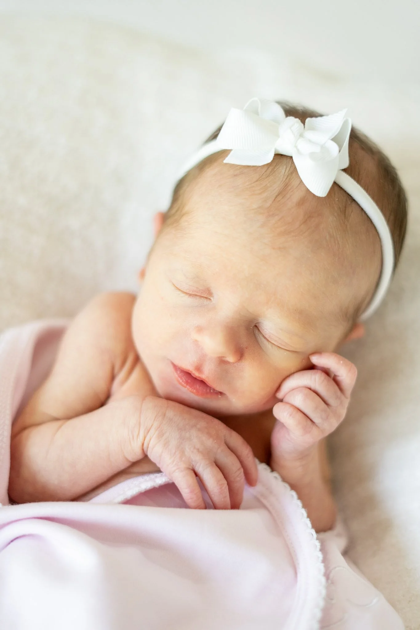 Close-up of a sleeping infant with a white bow on their head, resting peacefully on their hand.