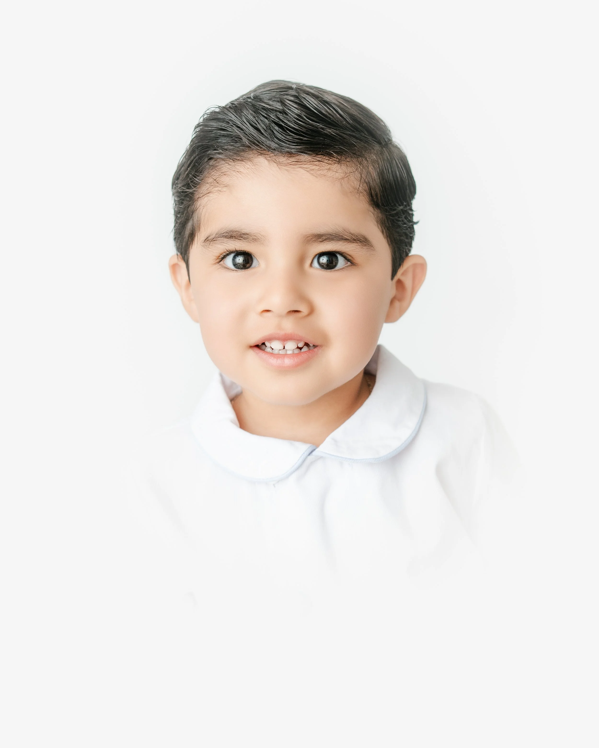 Close-up of a smiling young boy with black hair, blue eyes, and a white shirt against a plain white background.
