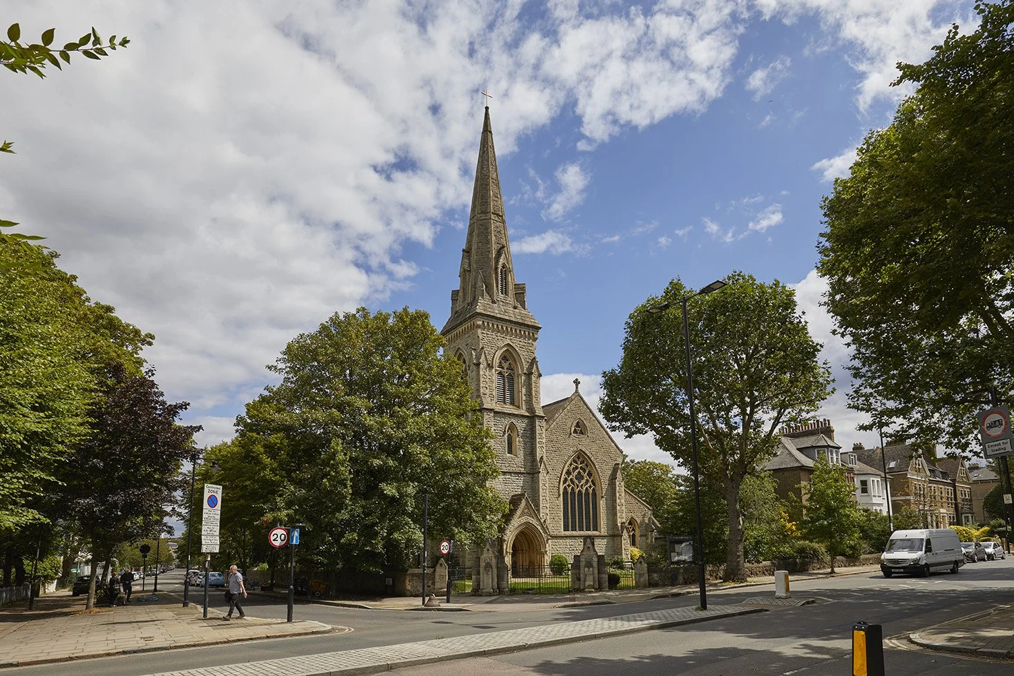 A stone church with a tall spire and Gothic windows in a small town, surrounded by trees and parked cars, on a partly cloudy day.