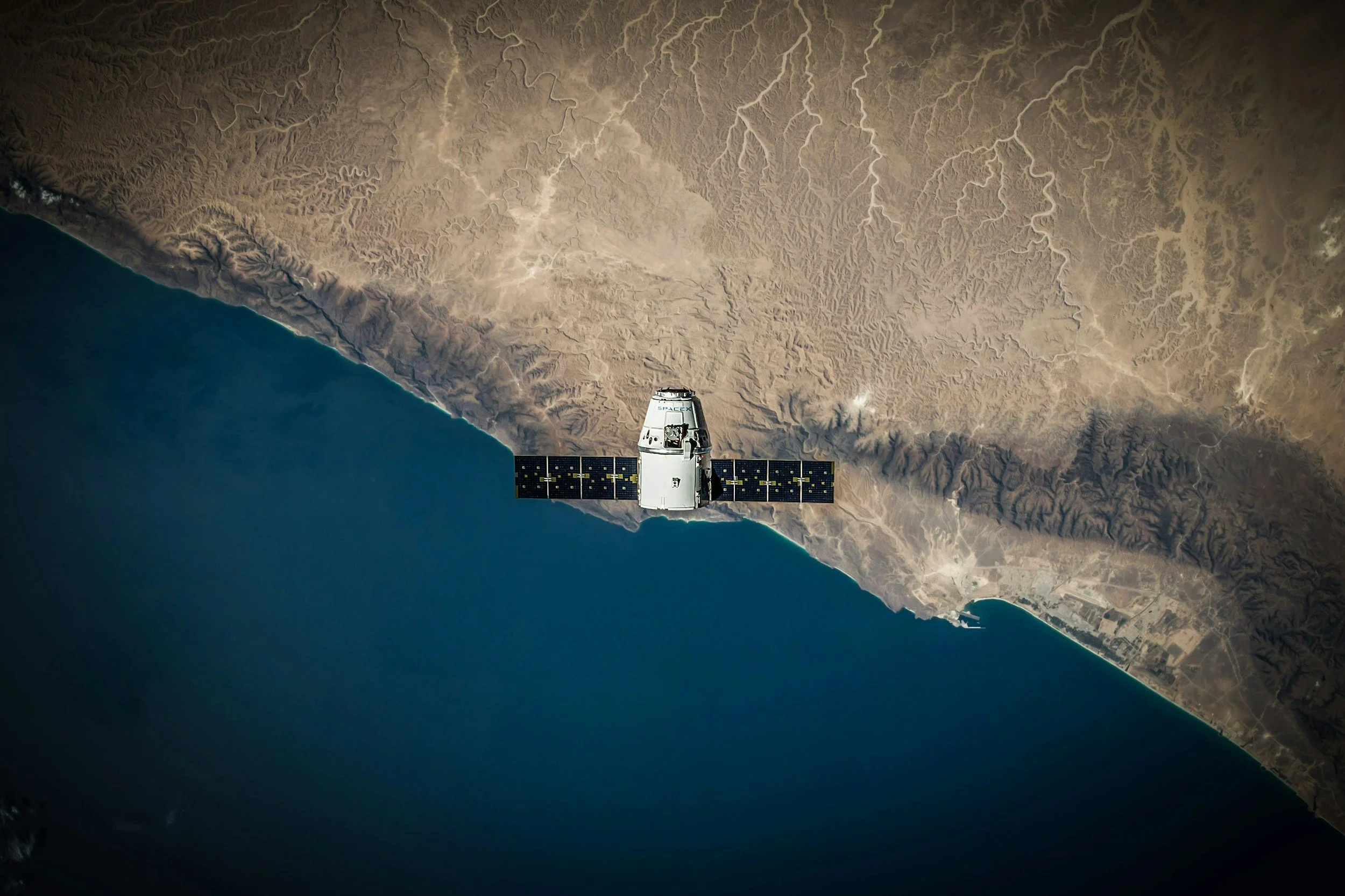 Satellite view of satellite in space above coastal terrain with mountains and desert, the ocean visible on the left.