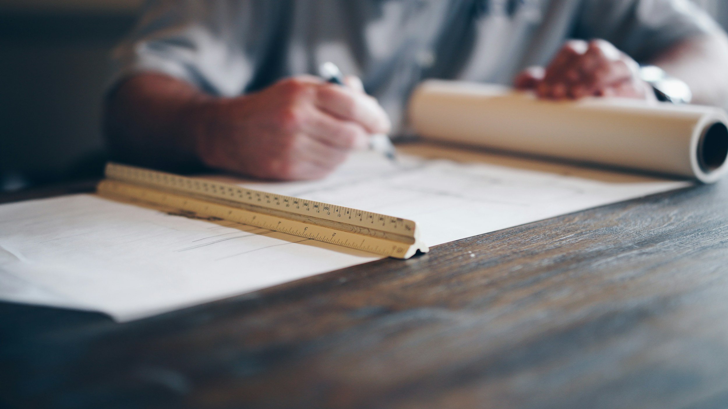 A person holds a pen over a rolled-up blueprint, with a wooden ruler and paper plans on a wooden table.