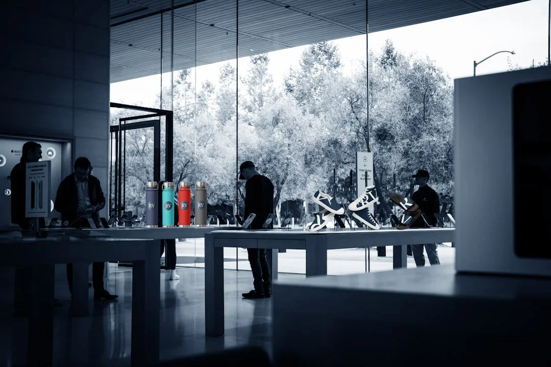 Inside an electronic store with large glass windows showcasing trees outside. Display tables hold colorful bottles, sneakers, and shoes. Customers browse, wearing face masks, and store staff assist.