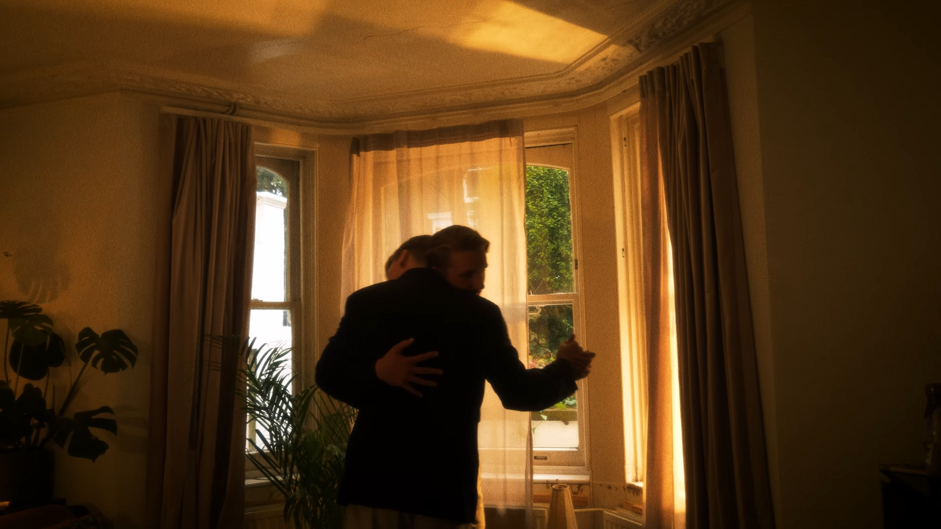 Two people hugging in a warmly lit room with large windows and beige curtains.
