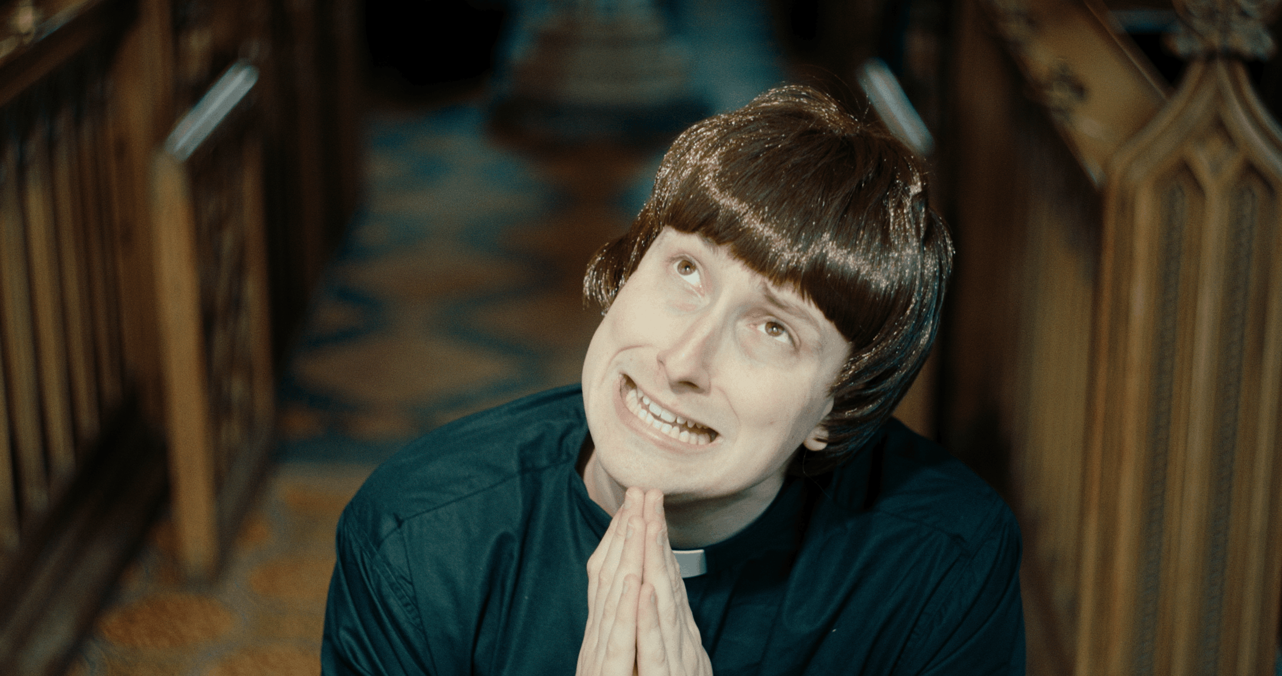 A woman with short brown hair kneeling on the ground with her hands pressed together in prayer, looking up with a smile inside a wooden church or chapel.