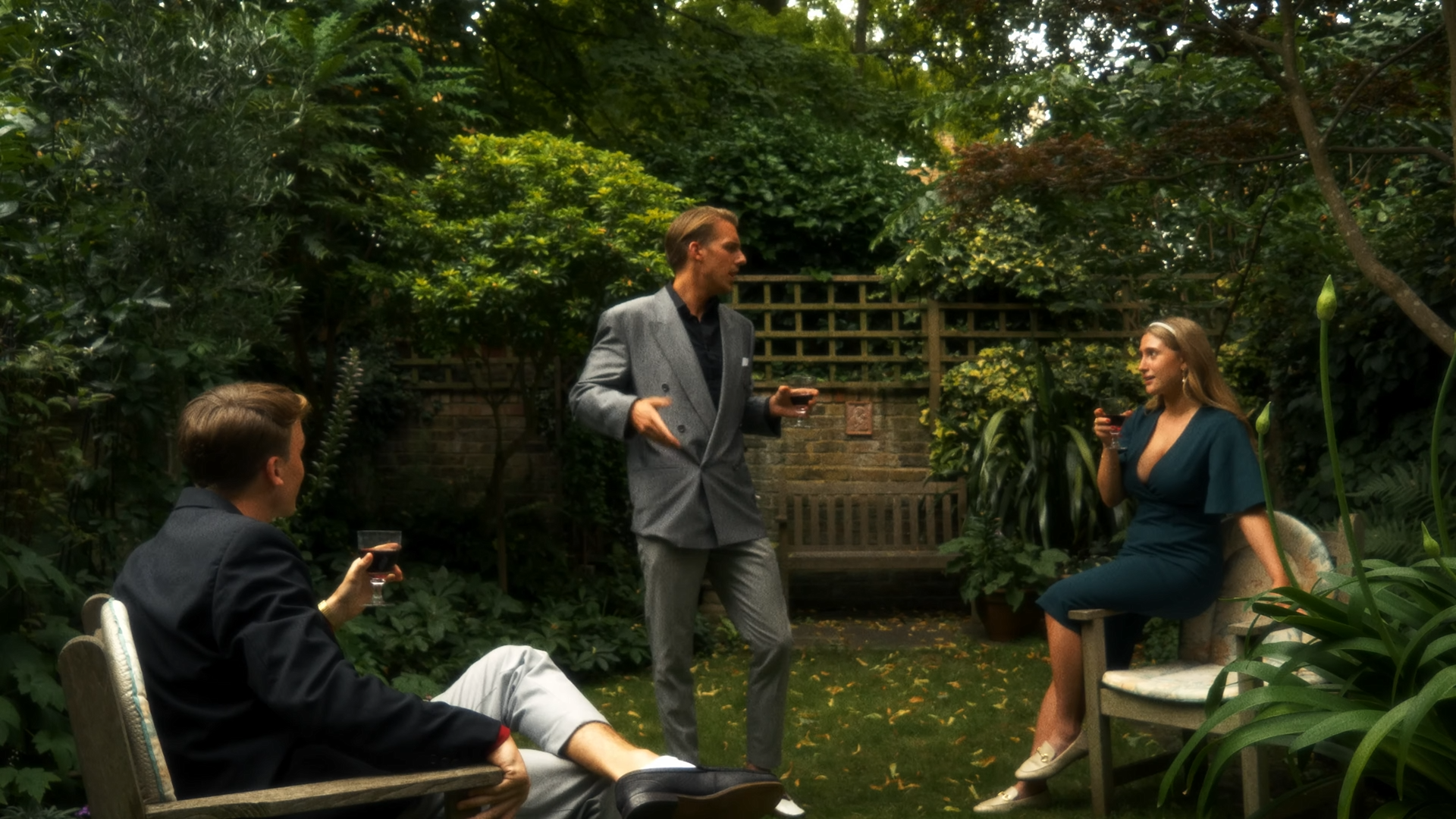 A group of four people in a lush garden having a conversation. One man is standing and speaking, while the others are seated, holding drinks.