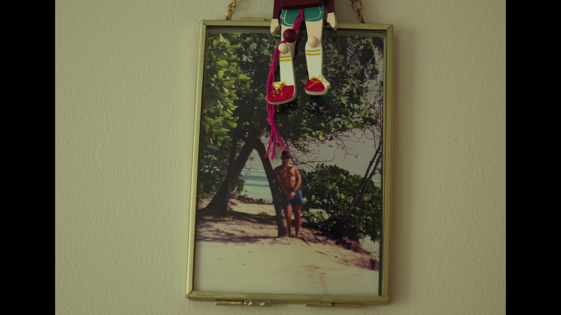A framed beach photograph with a man standing under a tree, hanging on a beige wall. A decorative hanging doll with red shoes and pink yarn is in front of the frame.
