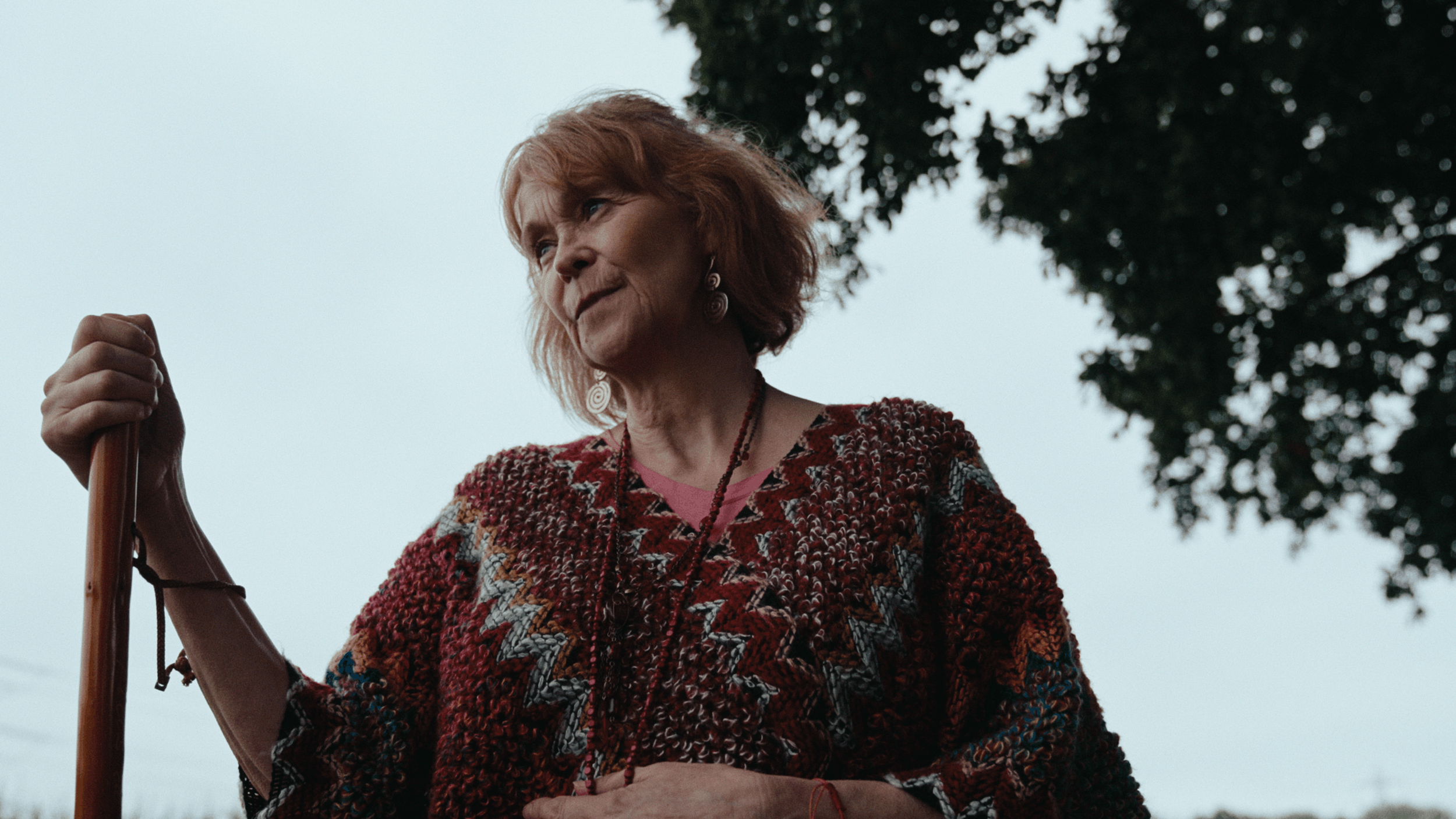 An elderly woman with red hair wearing a patterned red, black, and white sweater, holding a wooden walking stick outdoors near a tree with dark green leaves.