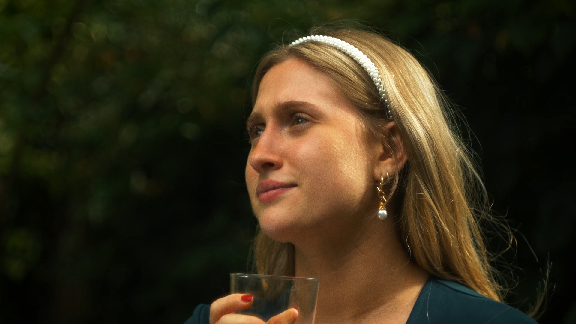 A young woman with blonde hair and light skin, wearing pearl earrings and a pearl headband, holding a glass, outdoors in front of a blurred green background.
