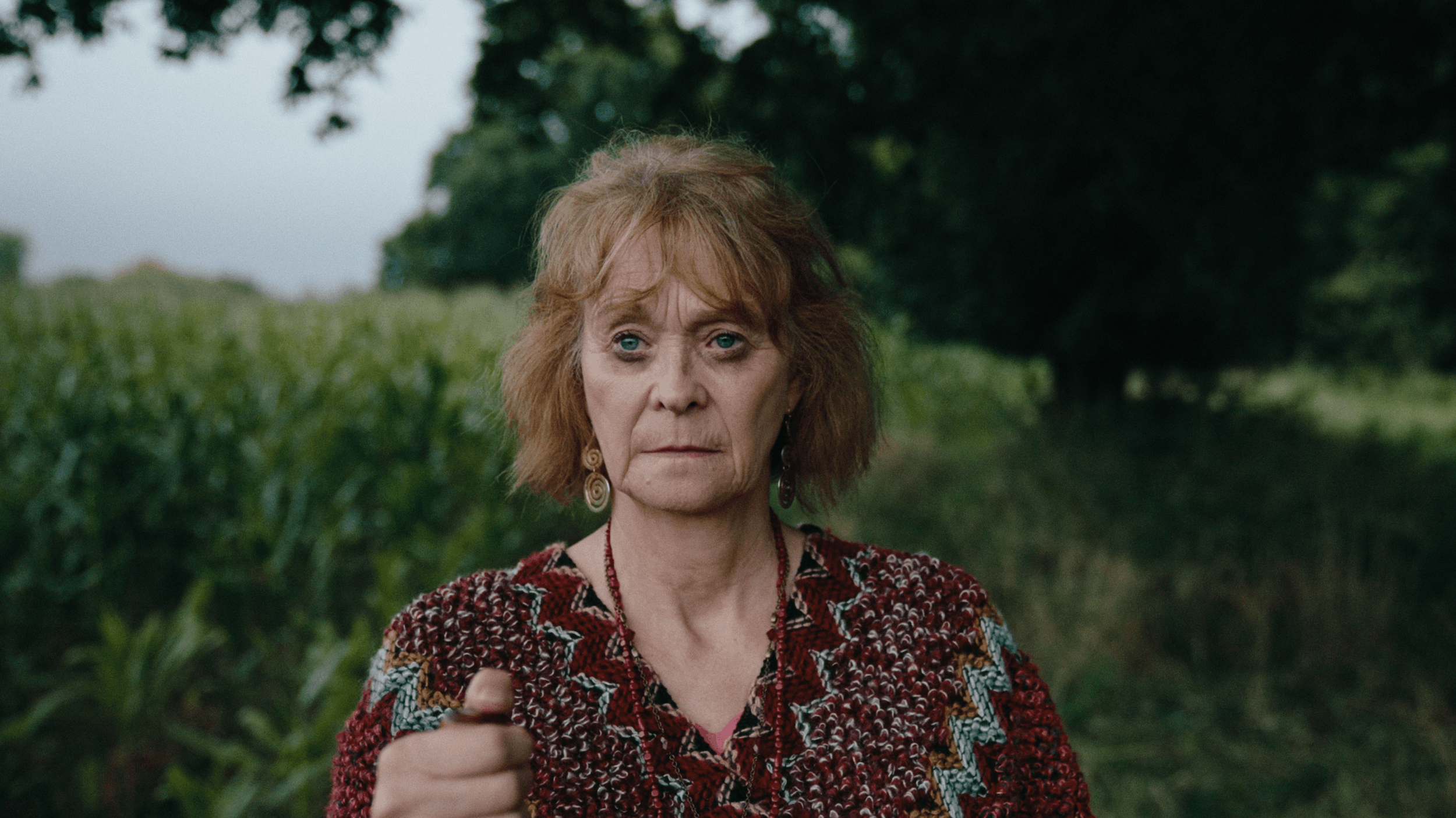 An older woman with red hair and blue eyes pointing towards the camera, standing outdoors with green trees and bushes in the background.