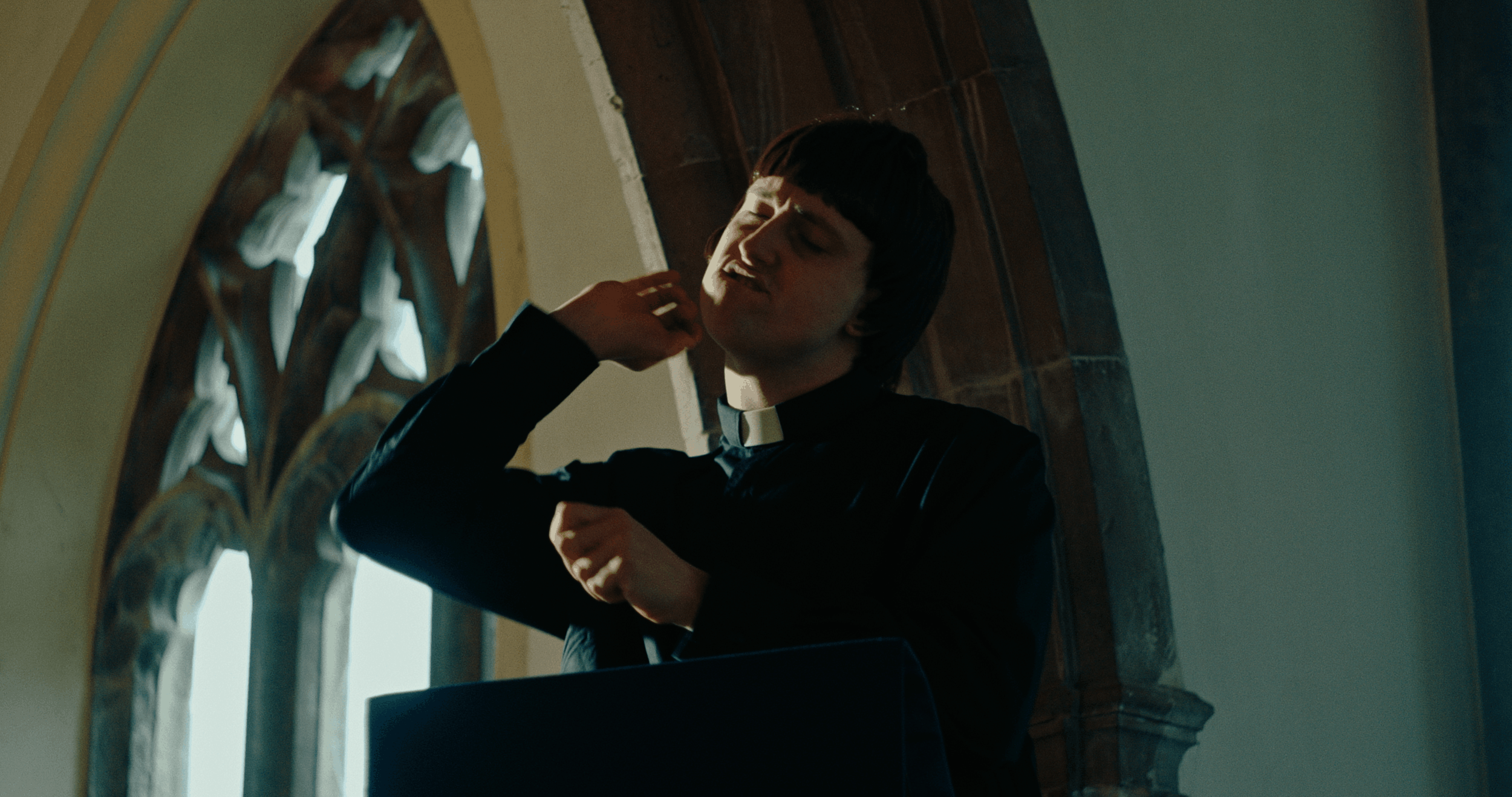 Young man dressed as a priest adjusting his collar inside a church, with arched windows and wooden paneling in the background.