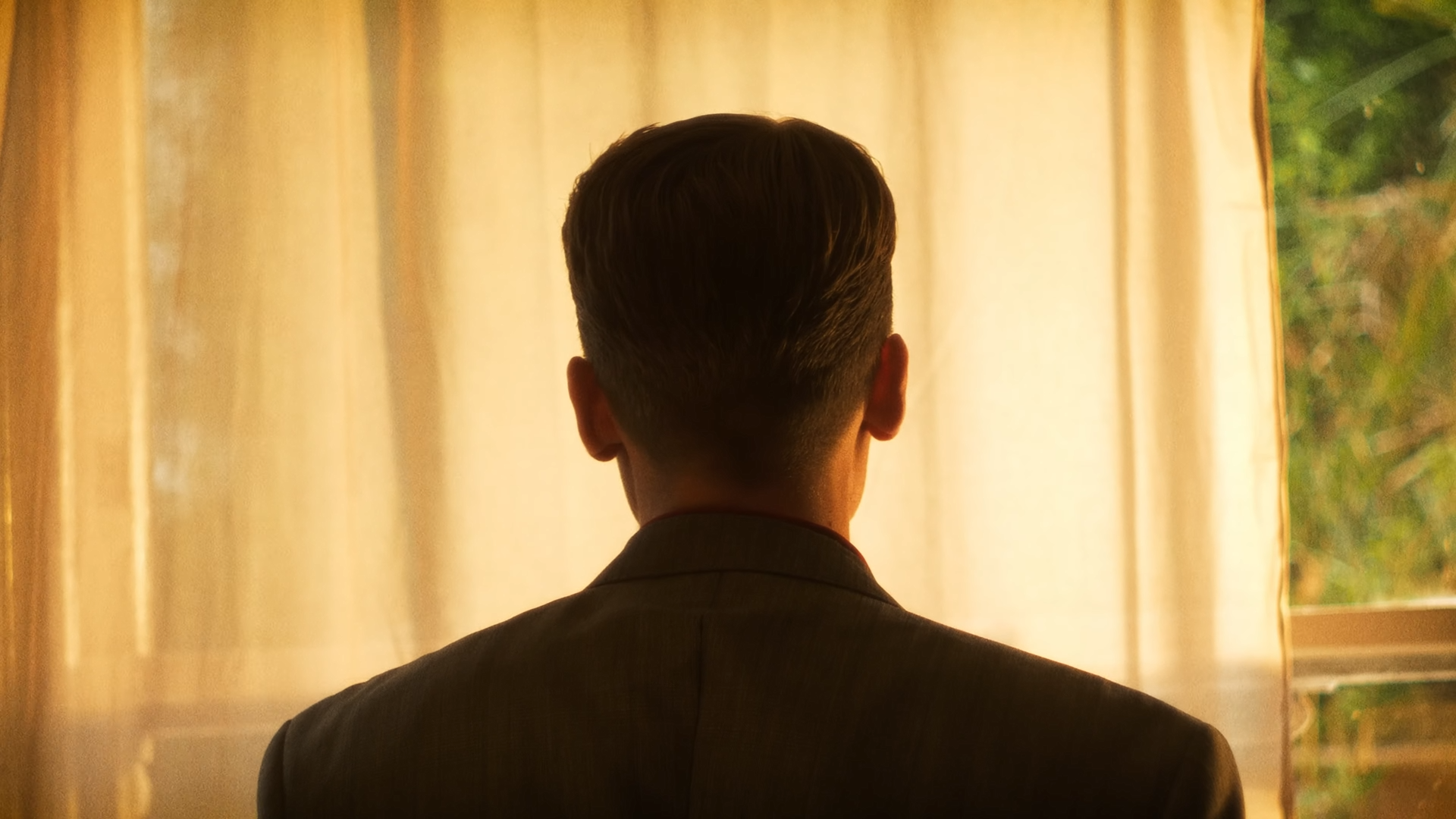 A man in a suit, seen from behind, looking out a window with yellow curtains during daytime.