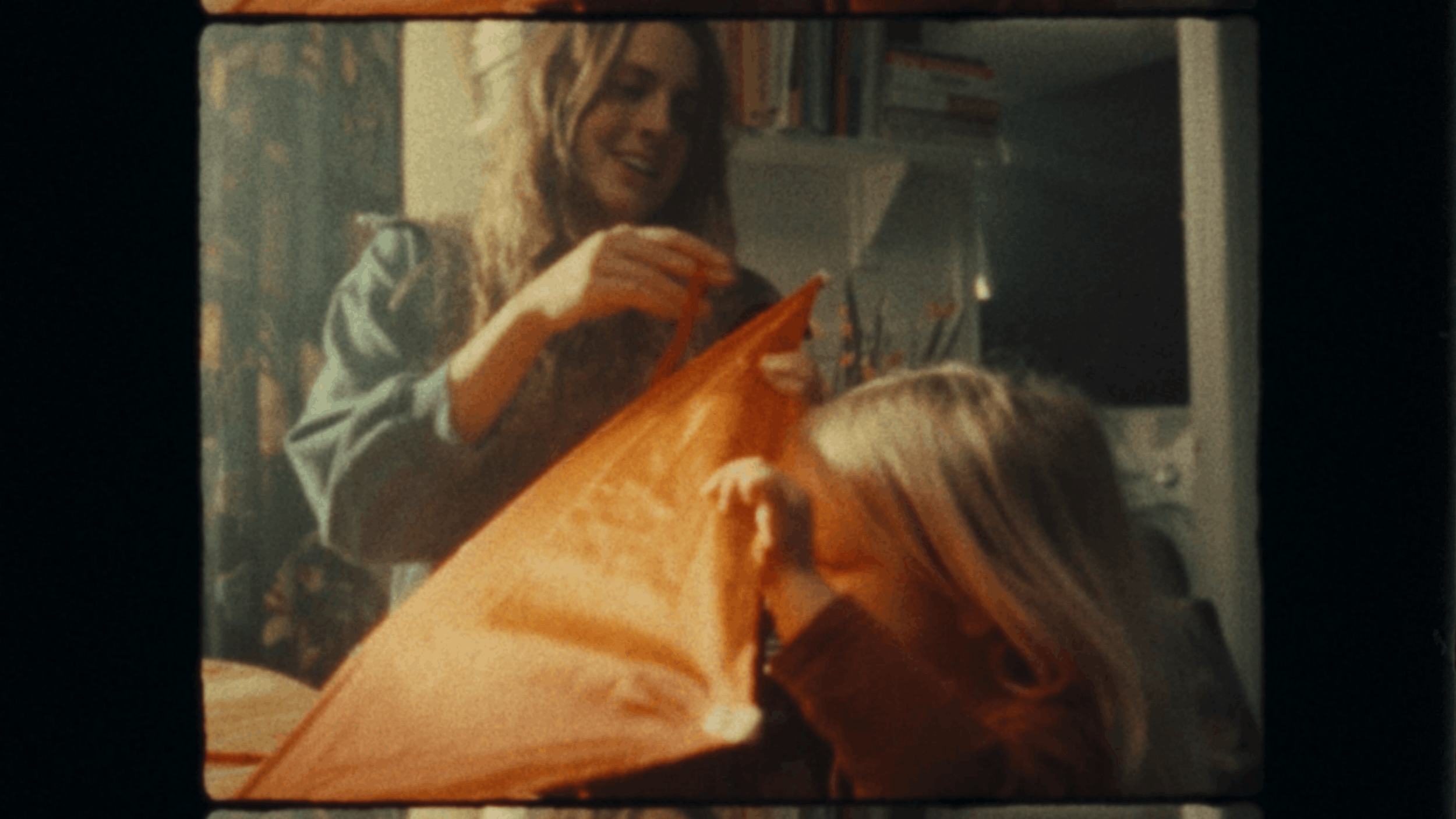 A woman with blonde hair smiling and holding a large orange shopping bag. She is in a room with bookshelves and appears to be happy.
