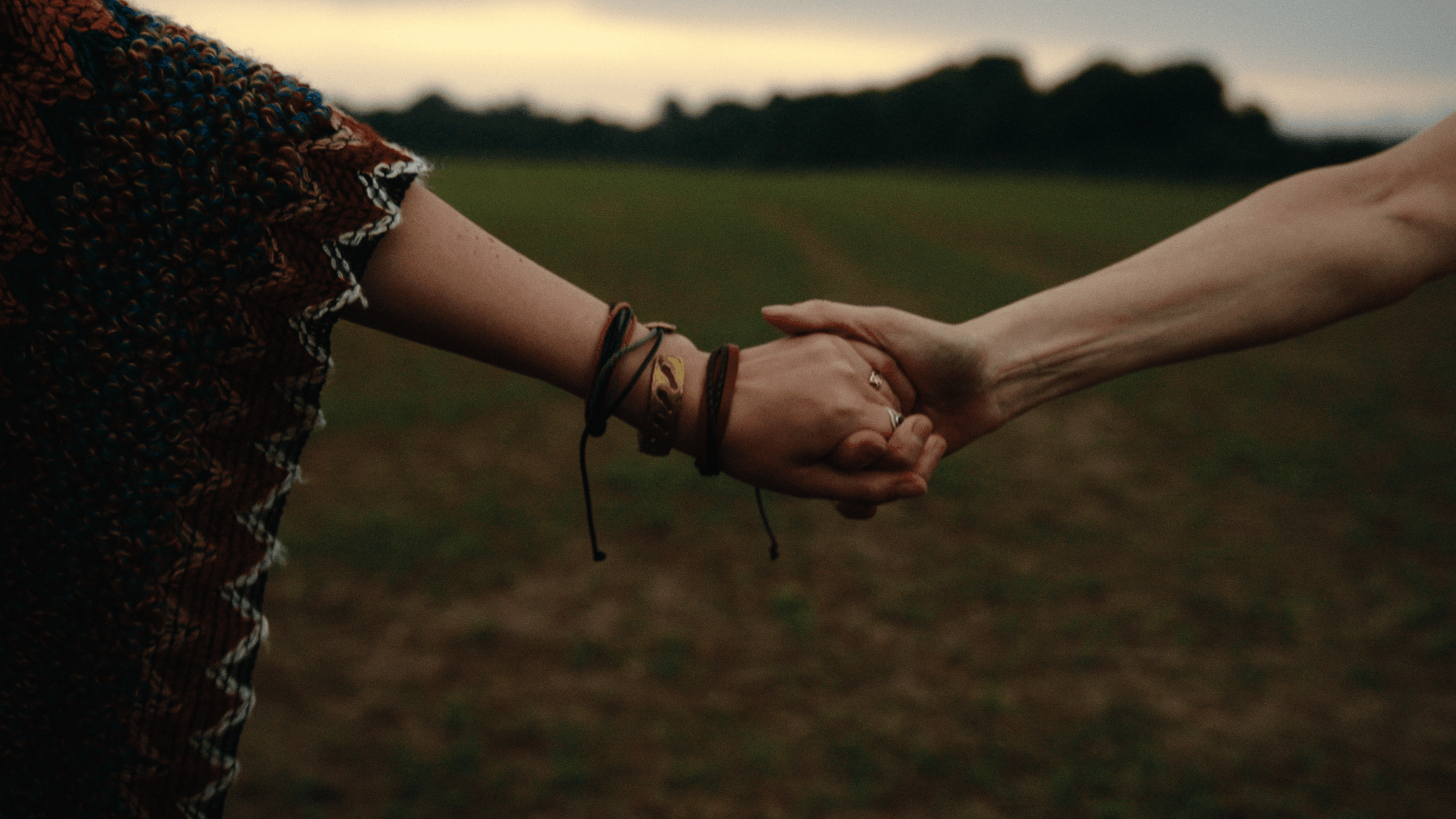 Two people holding hands outdoors during sunset or dusk, with a blurred grassy field and hills in the background.
