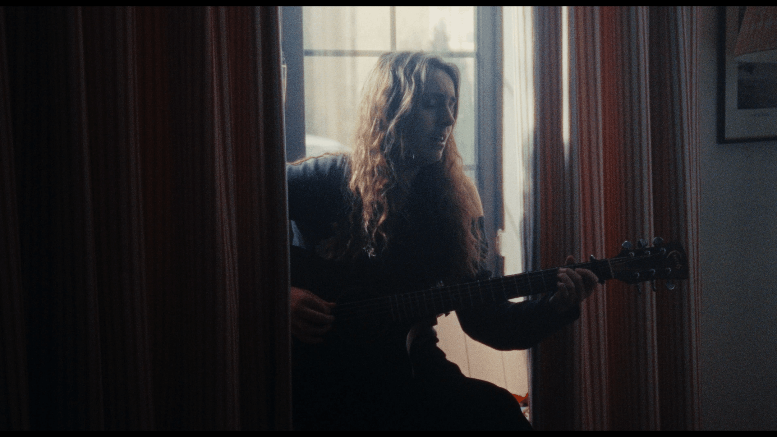 A woman with curly hair playing an acoustic guitar in a room with curtains and natural light.