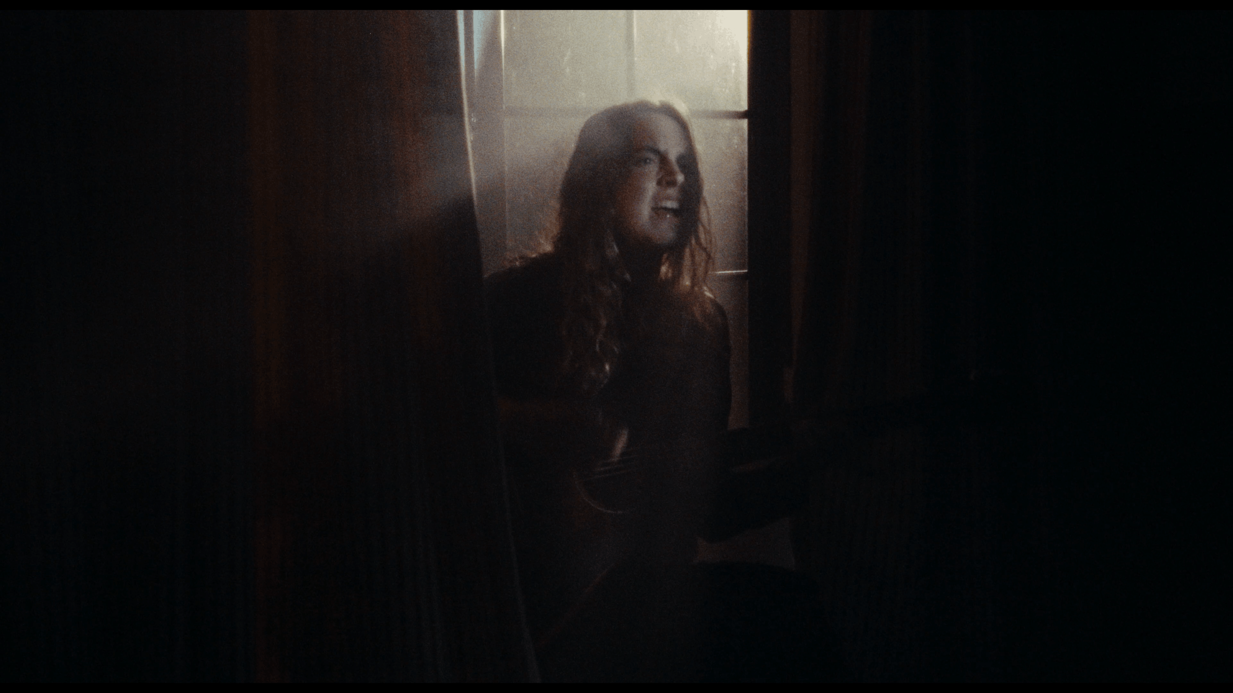 A woman with long, wavy hair, smiling and looking to her right, sitting in a corner near a window with sunlight coming through, in a dimly lit room.