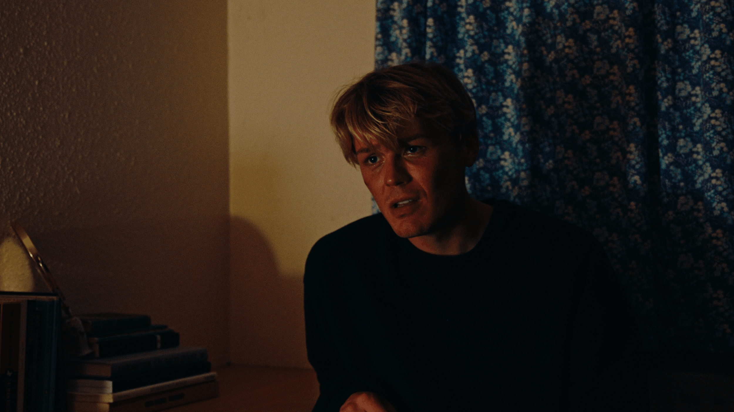 A young man with light skin and brown hair styled with a blonde streak, sitting on a chair in a dimly lit room. He appears distressed or thoughtful, with a somber expression on his face. Behind him, there's a blue floral curtain, and to the left, part of a bookshelf with books and a keyboard are visible.