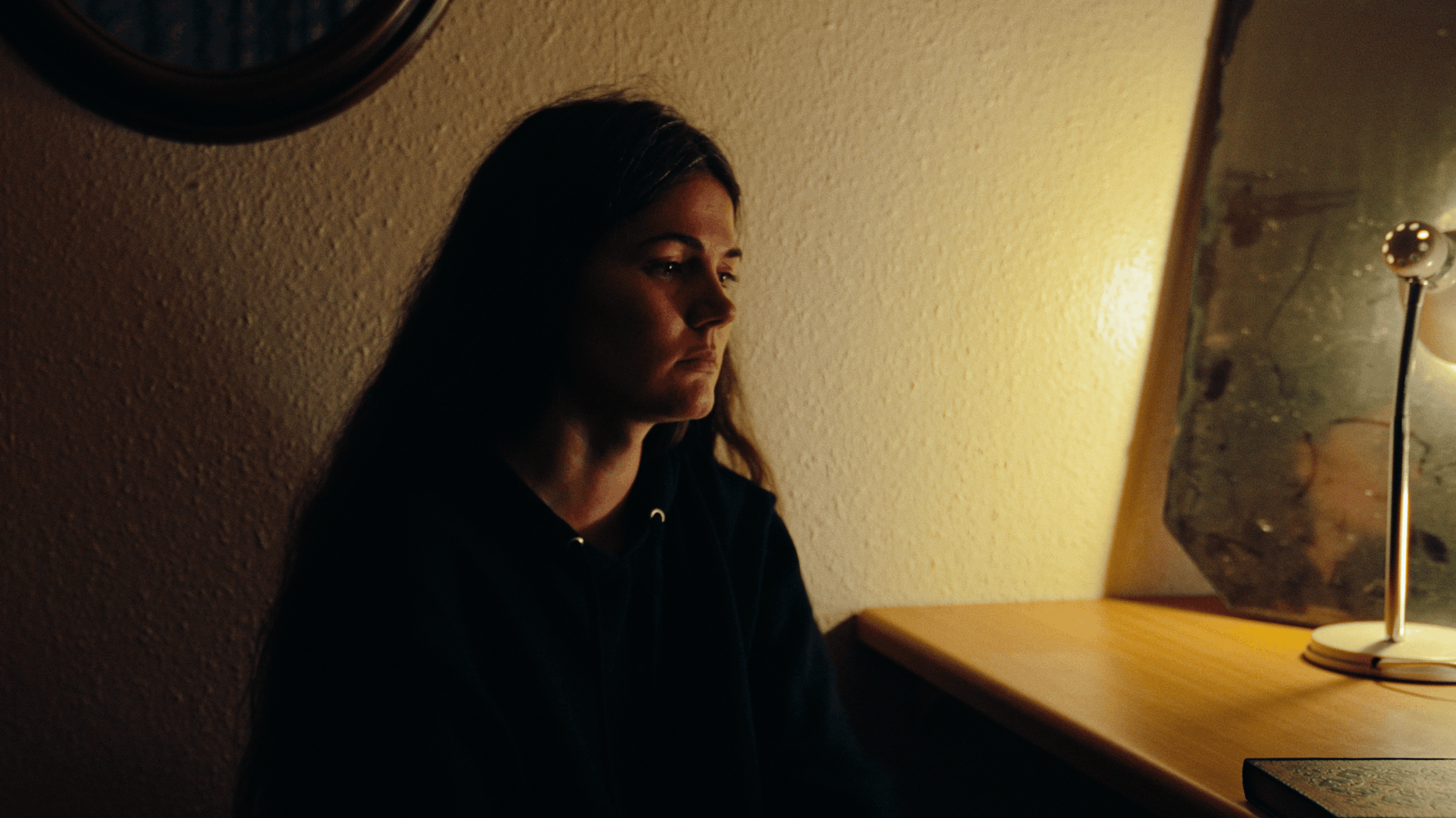 A young woman with long dark hair sitting at a wooden desk, looking at a mirror with a sad or contemplative expression, illuminated by warm yellow lighting.