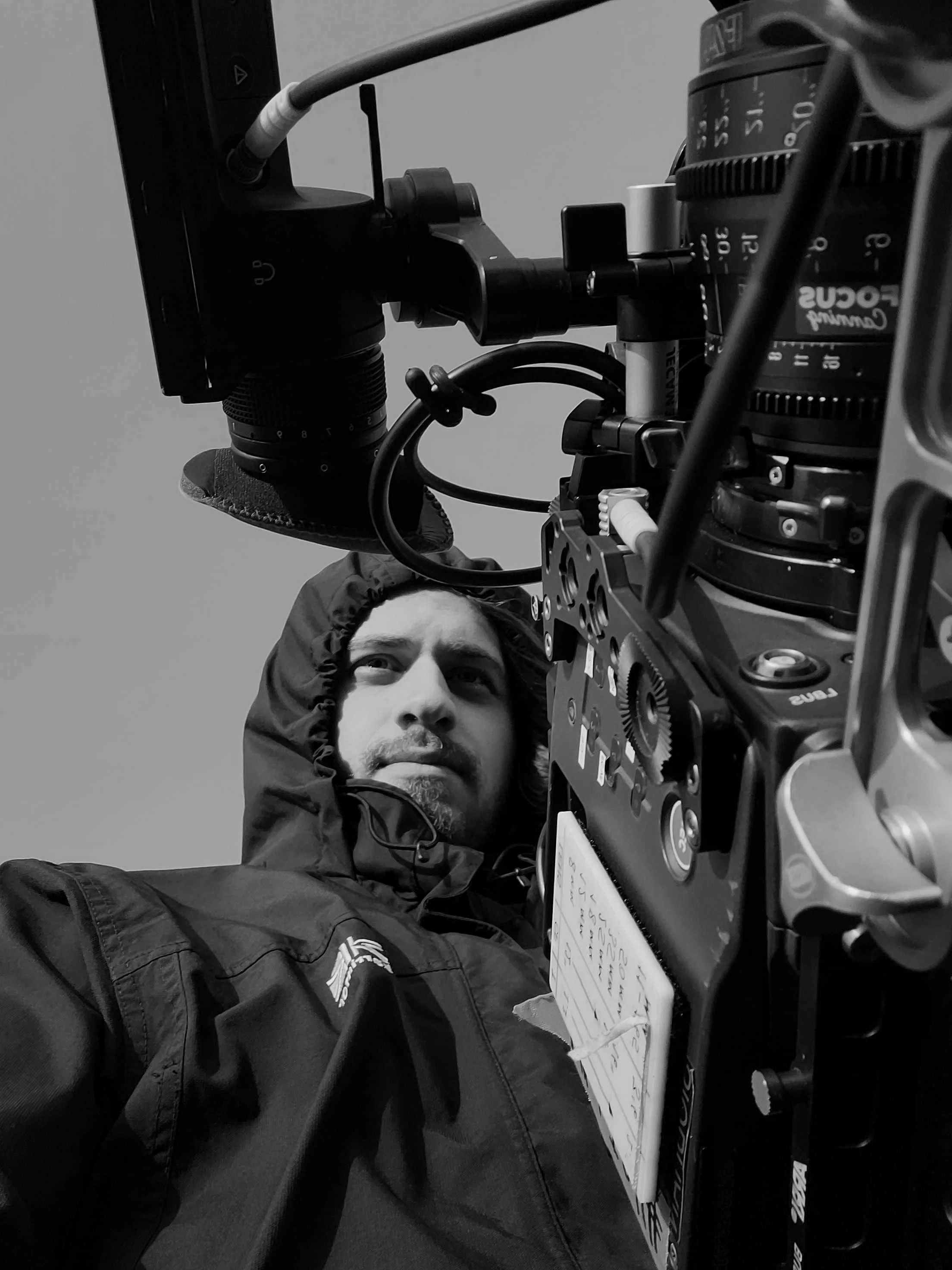 A man wearing a hooded jacket looking through a professional camera with various dials and controls, with a handwritten note attached to the camera.