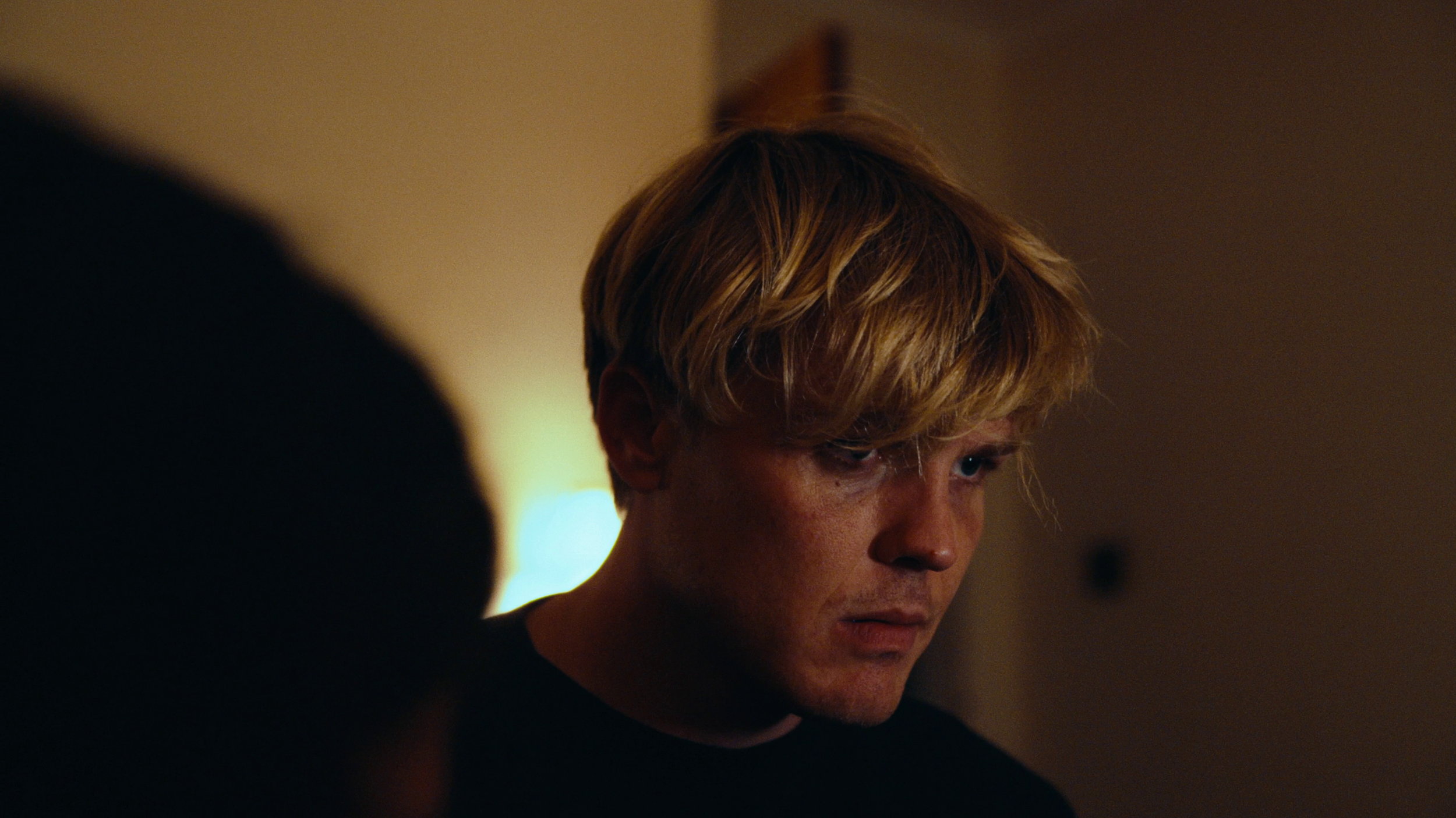 A young man with blonde hair looking down in a dimly lit room.