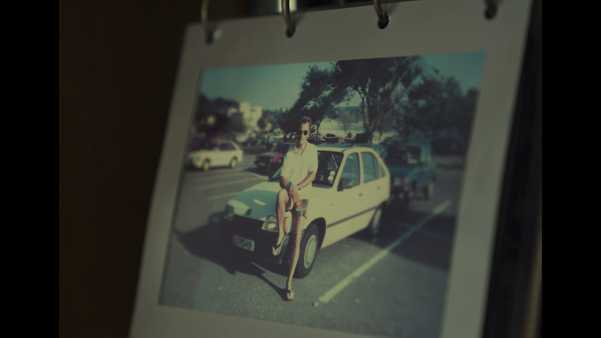 A printed photograph on a stand showing a woman sitting on the hood of a small white car in a parking lot with other cars and trees in the background.