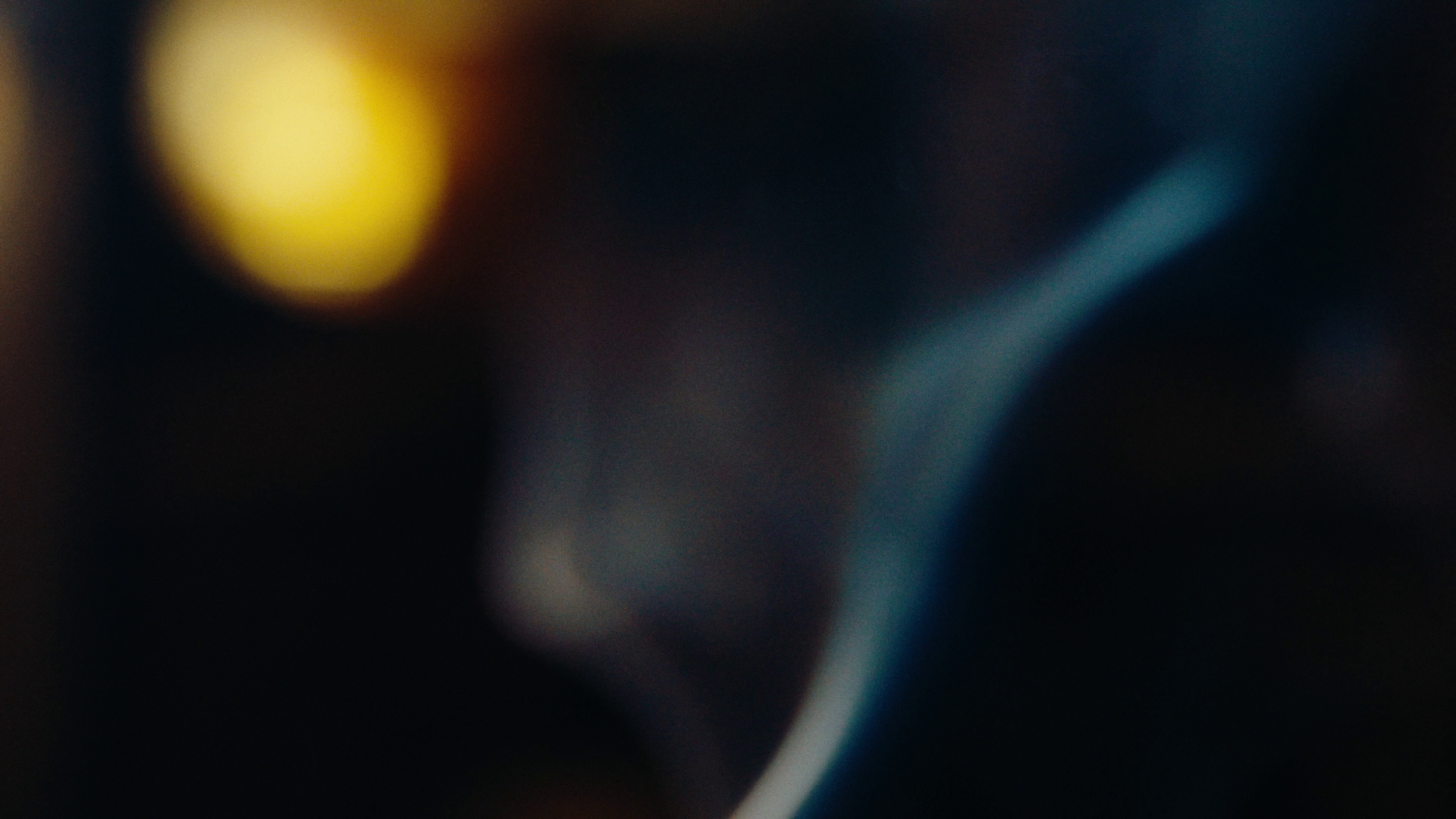 An abstract, blurry photograph with dark tones, featuring a prominent yellow shape on the left and a faint blue curve on the right.