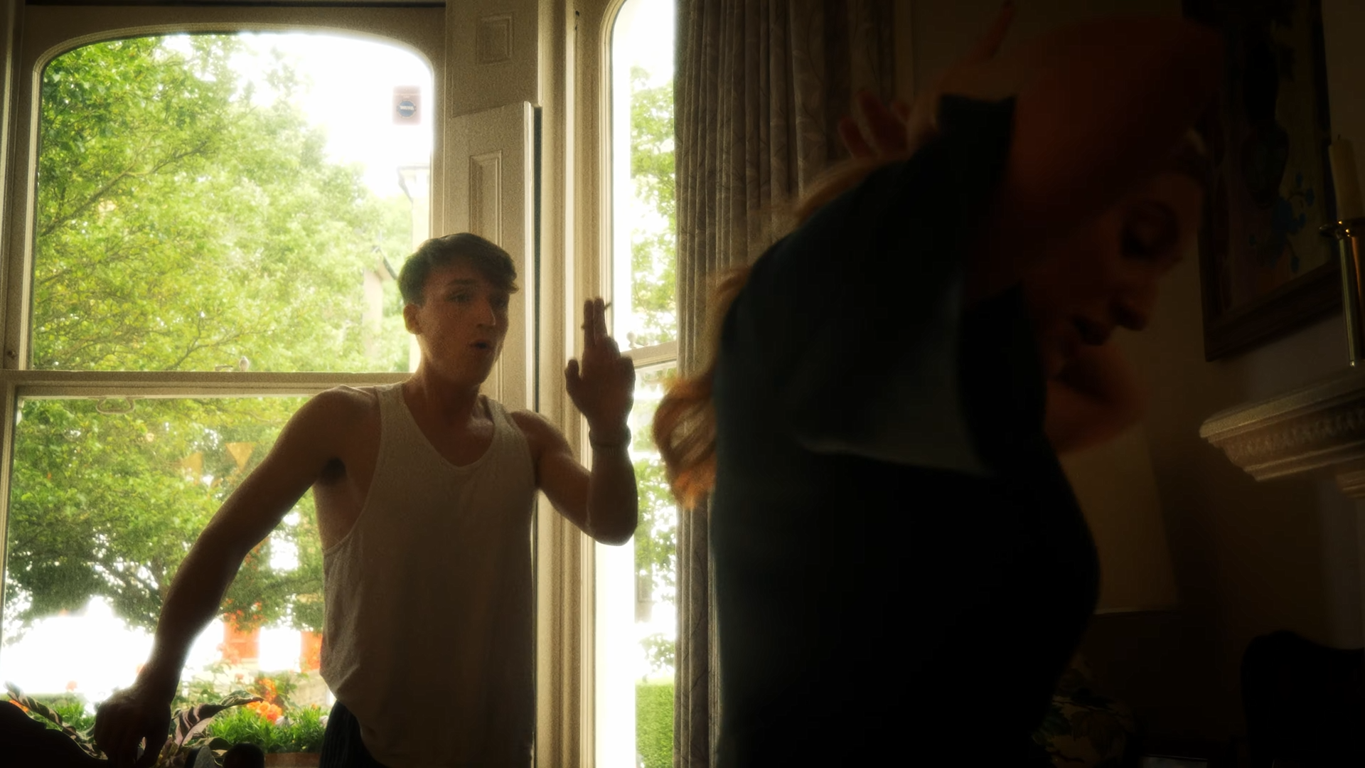 A young man in a white sleeveless shirt raising his left hand as if making a gesture, standing by a large window with daylight and green trees outside, while a young woman with long hair, wearing a dark top, is in the foreground with her face turned downward in a dimly lit room.