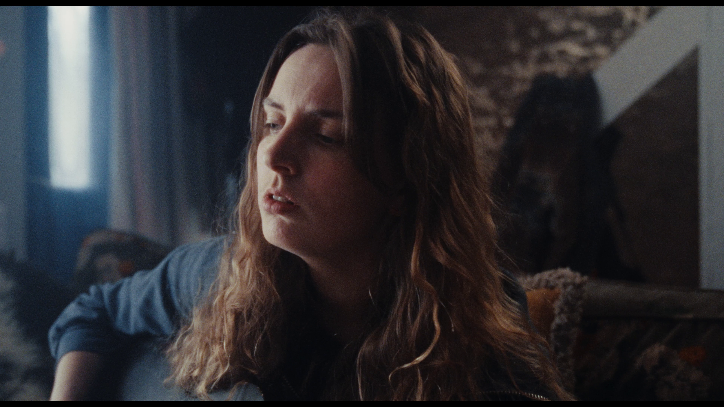 A woman with long, wavy auburn hair, wearing a blue shirt, appears to be in a moment of contemplation or sadness indoors with a brick wall in the background.