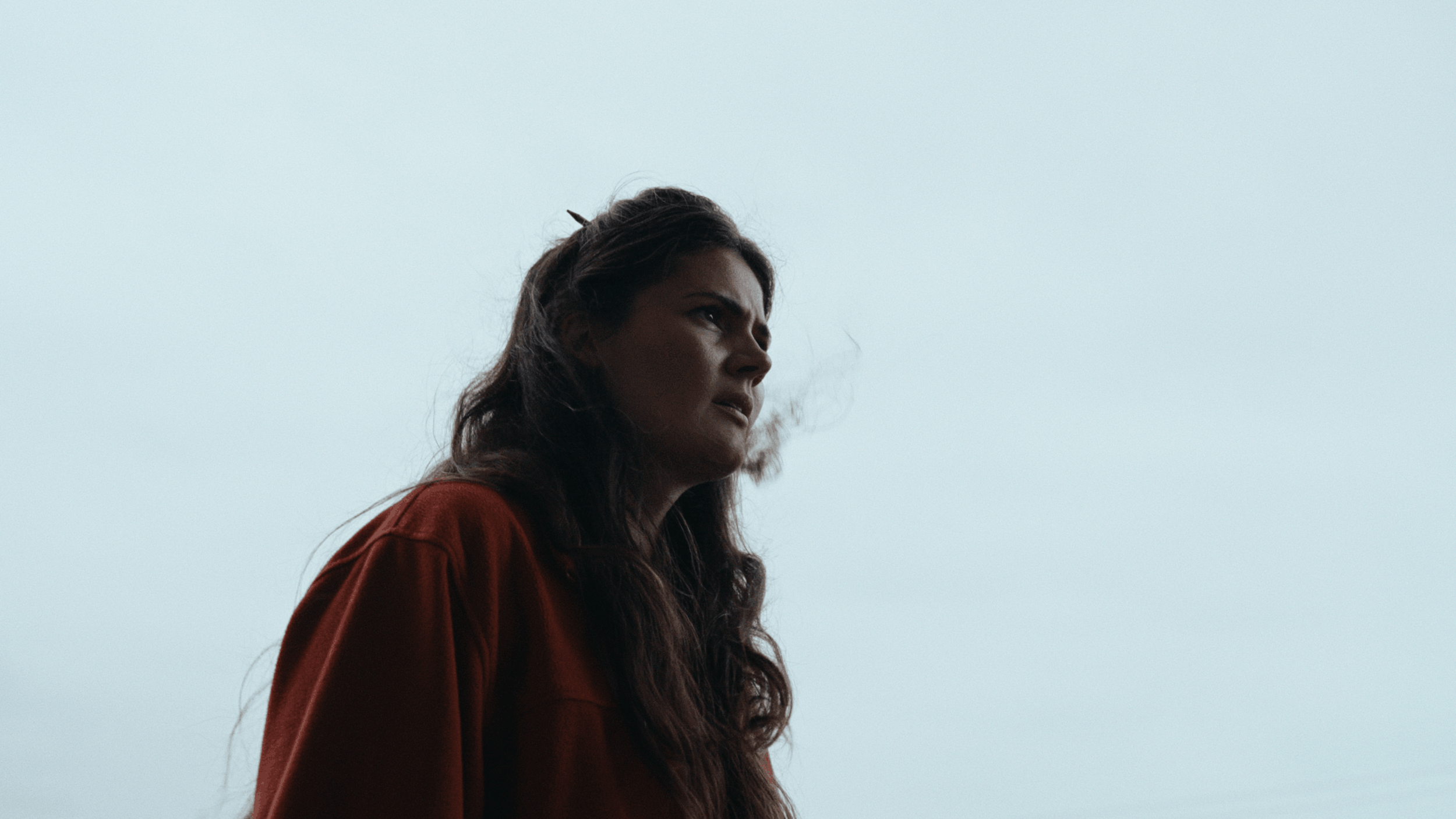 A woman with long, dark hair looking to the side against a gray sky, wearing a red jacket.