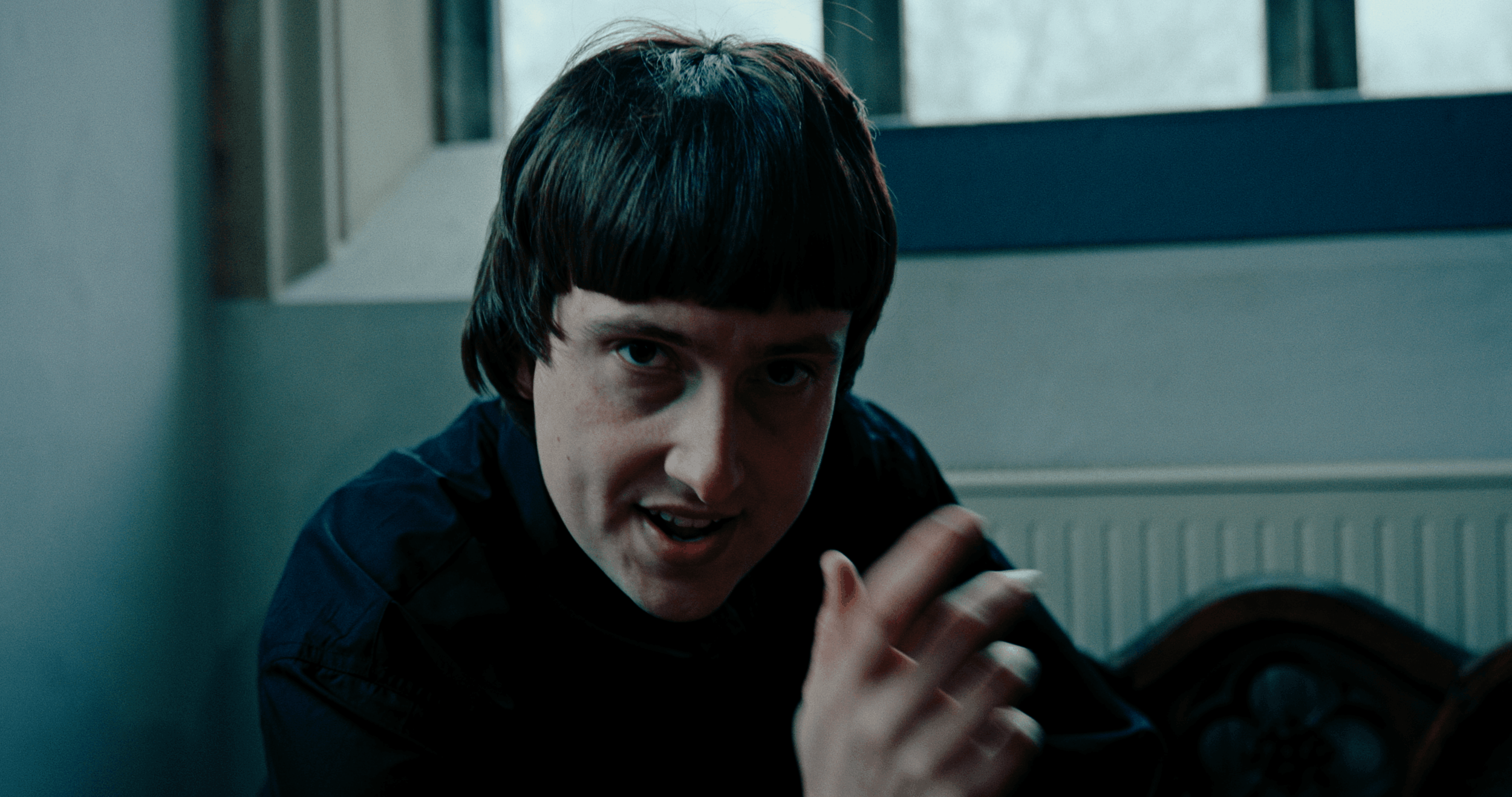 A person with short dark hair and a dark shirt, looking up and speaking, indoors near a radiator and window.
