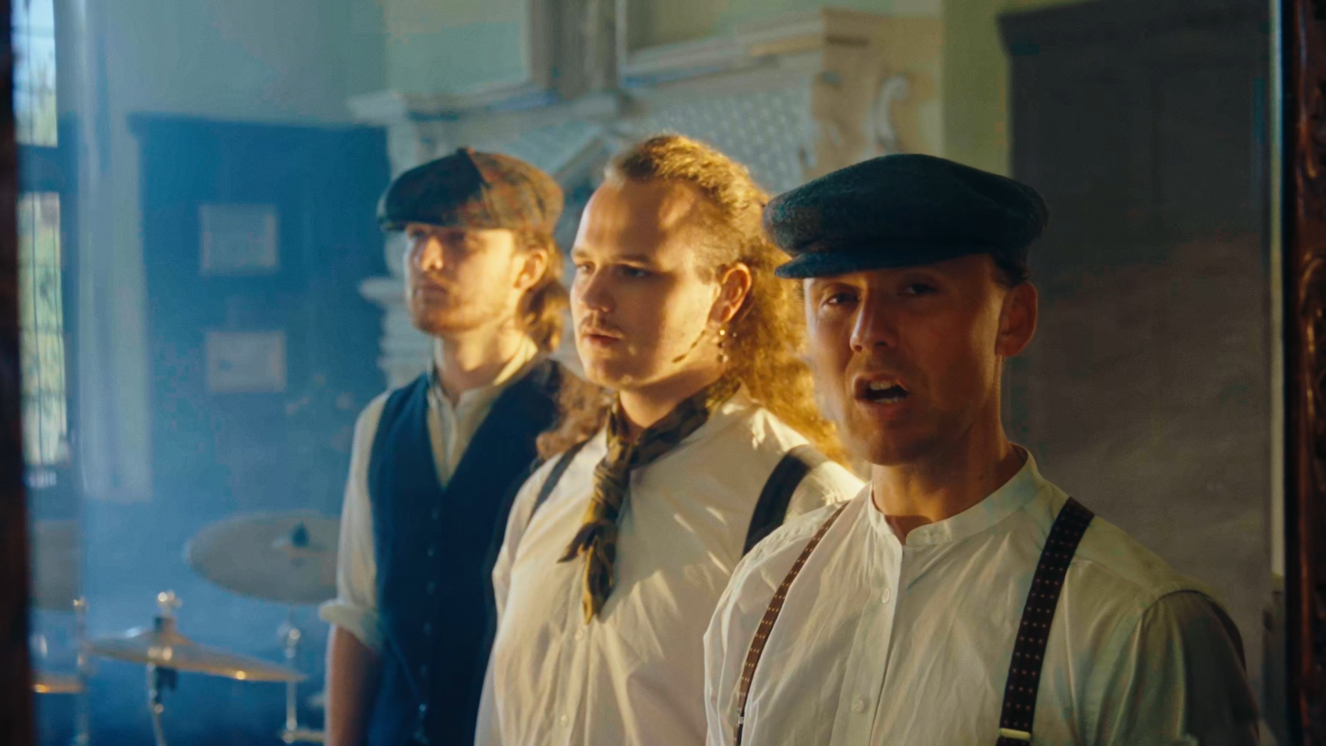 Three men with vintage clothing look through a glass window; one is speaking and has suspenders, the second has curly hair, and the third wears a plaid cap.