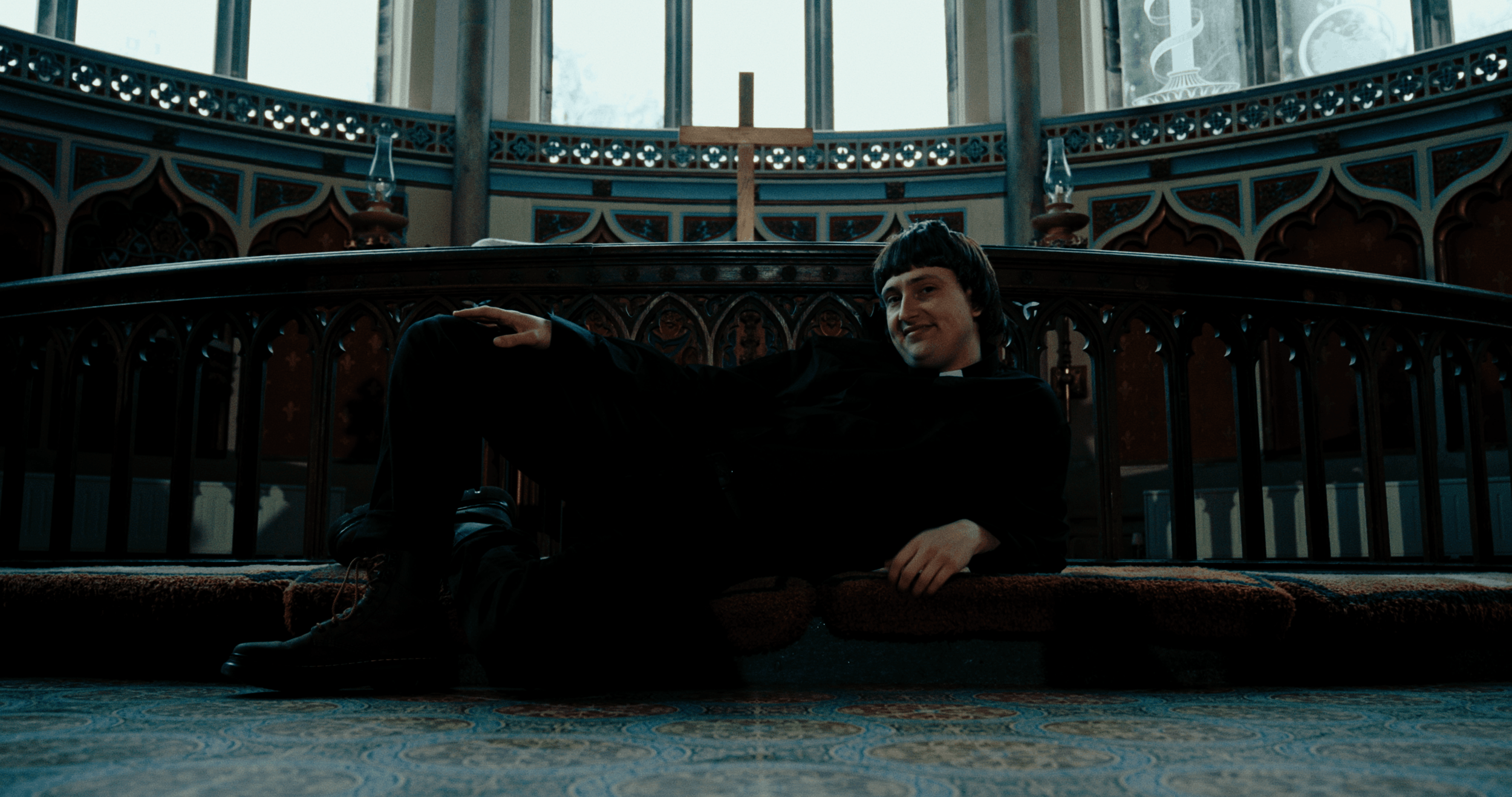 Young man in a black outfit sitting and reclining on the floor of a church or chapel with intricate woodwork, a cross, and stained glass windows in the background.