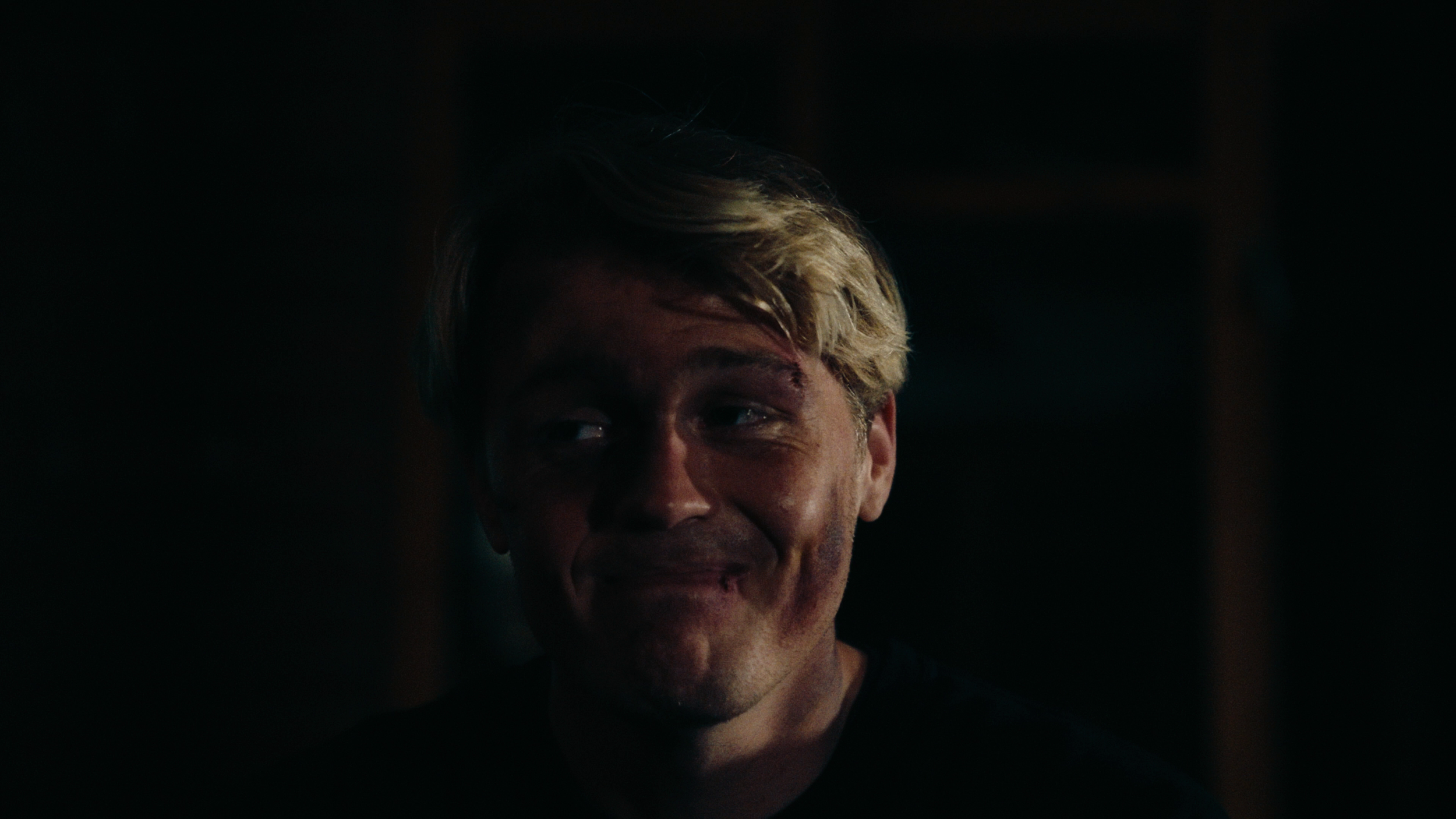 Close-up of a man with blond hair and a smile, partially visible in a dark setting.