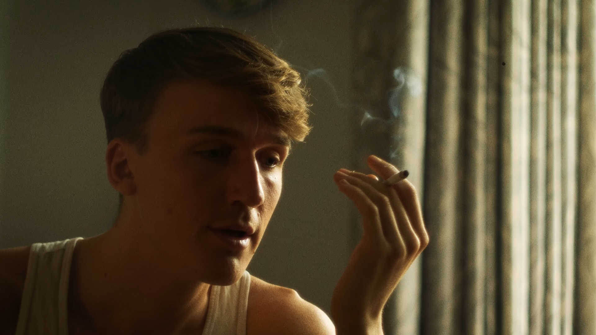 A young man with short, wavy hair is smoking a cigarette indoors near a window with closed curtains. The lighting is dim and warm.