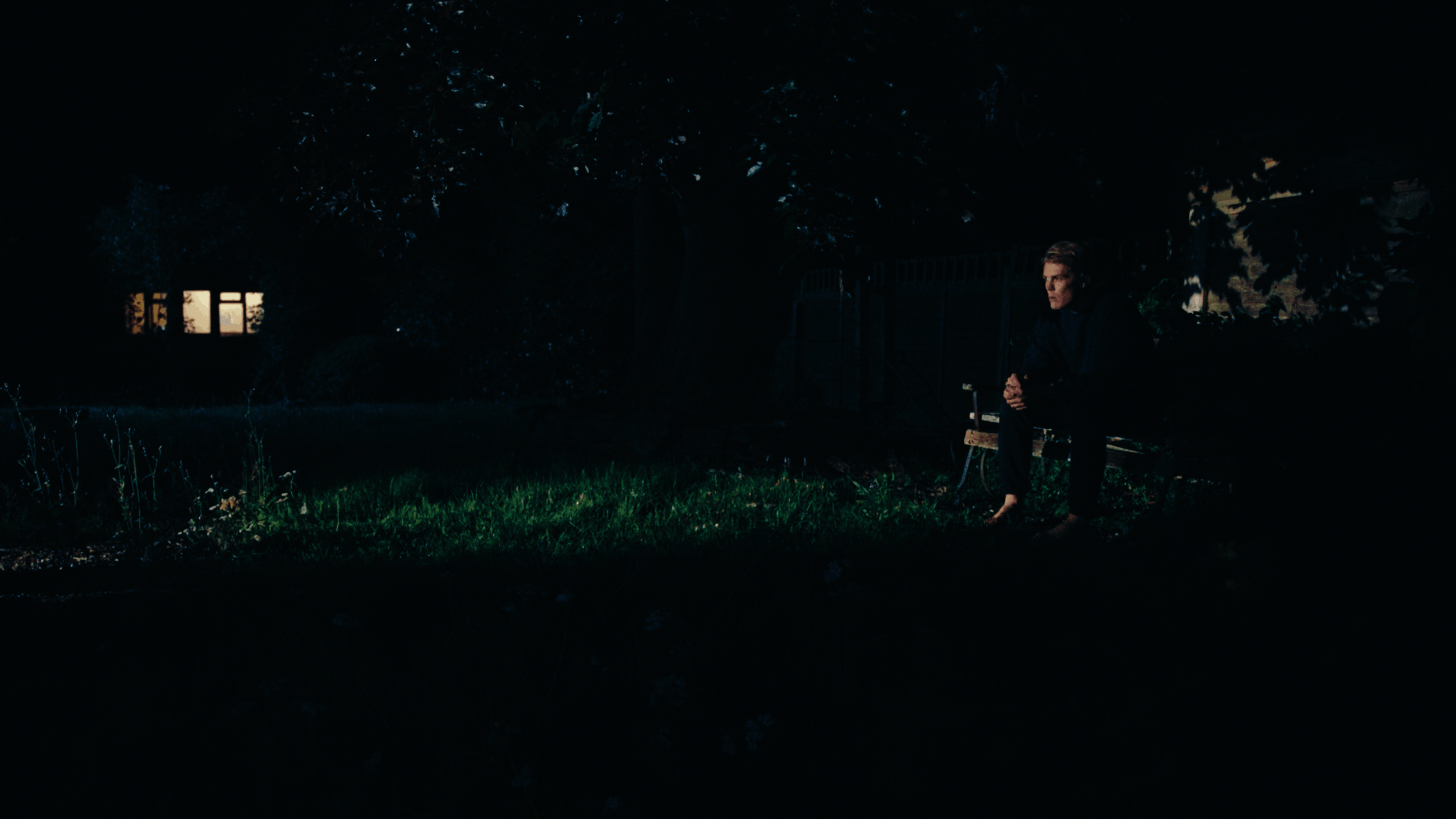 A man sitting on a park bench at night, dressed in black, with a dark background and a house with lit windows in the distance.