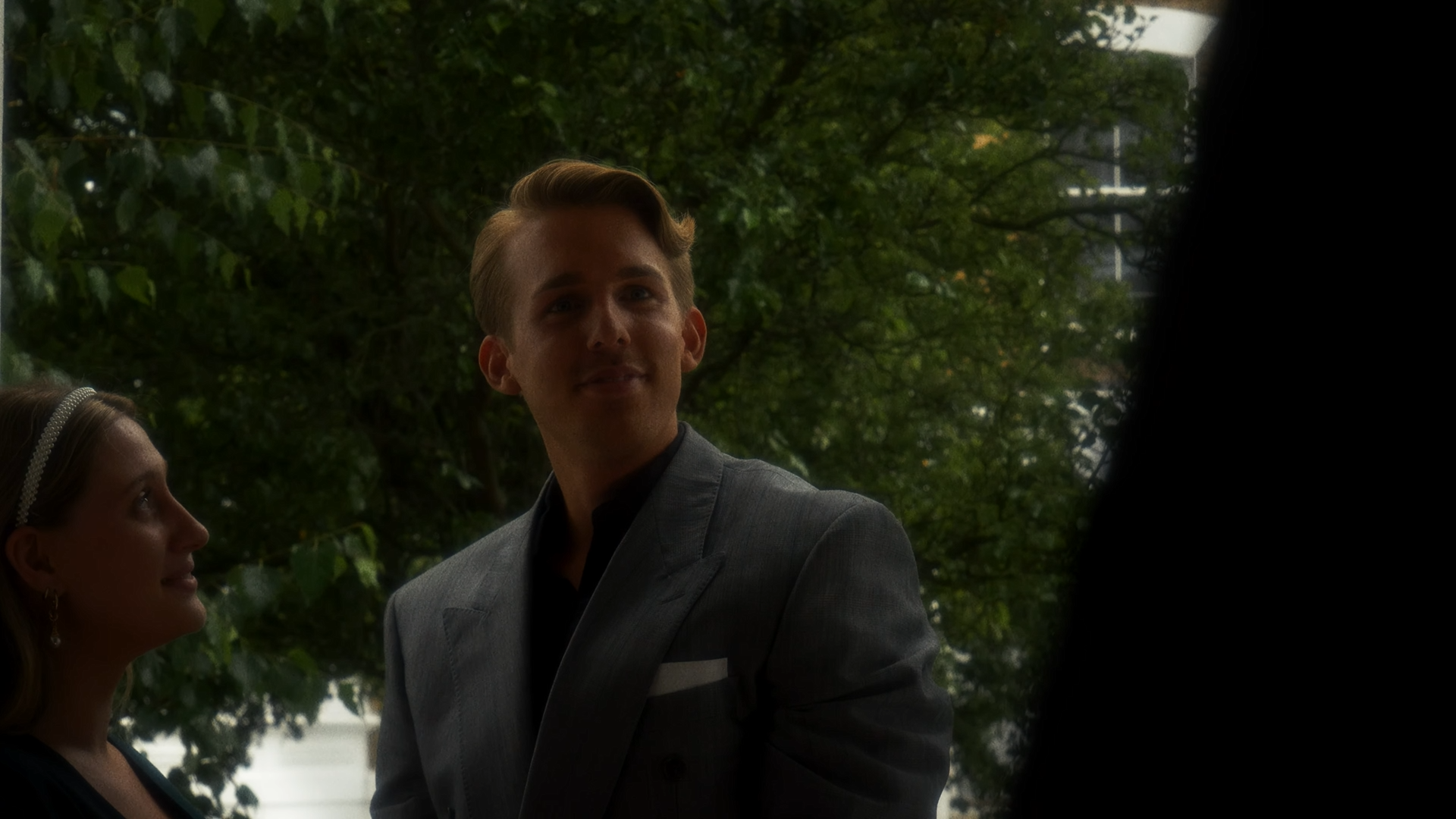 A young man wearing a grey suit and black shirt, standing outdoors with trees and greenery in the background. Partially visible is a young woman wearing earrings and a headband, looking at him.