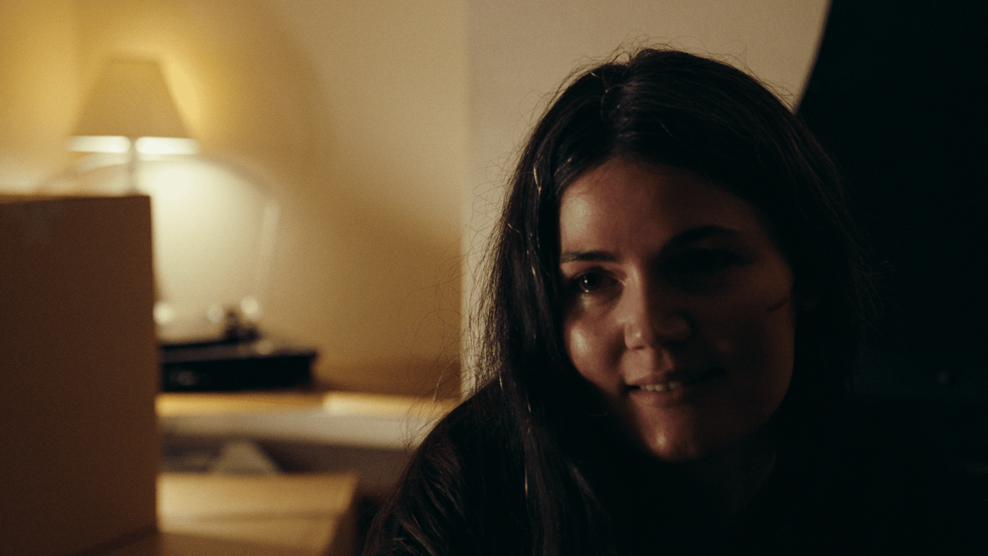 A woman with long dark hair smiling softly, sitting in a dimly lit room with a beige wall and a nightstand with a lamp in the background.
