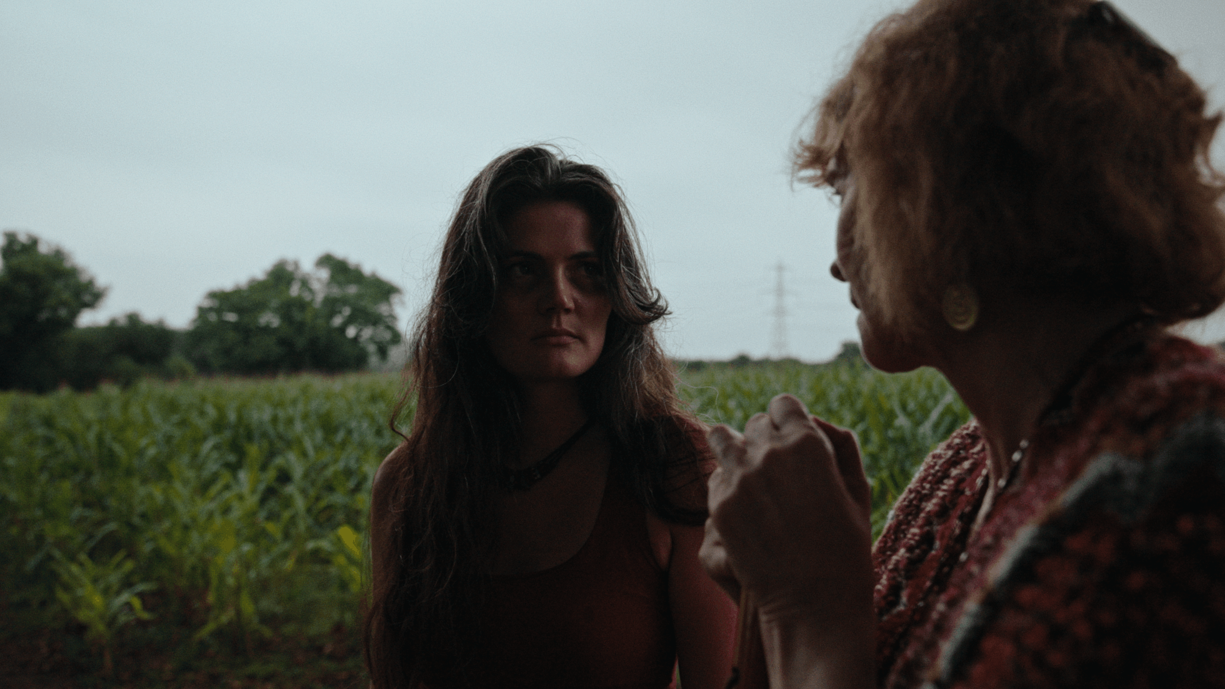 A young woman and an elderly woman having a conversation outdoors in a field with greenery and trees in the background.