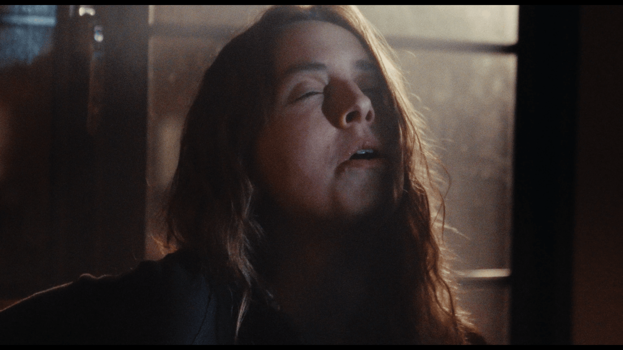 A woman with wavy hair and closed eyes appears to be in a moment of emotional intensity, with sunlight streaming in from the side.