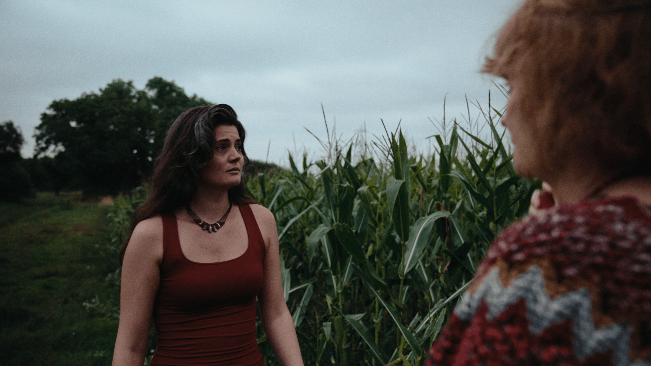 Two women standing outdoors in a field of tall crops, facing each other on a cloudy day.