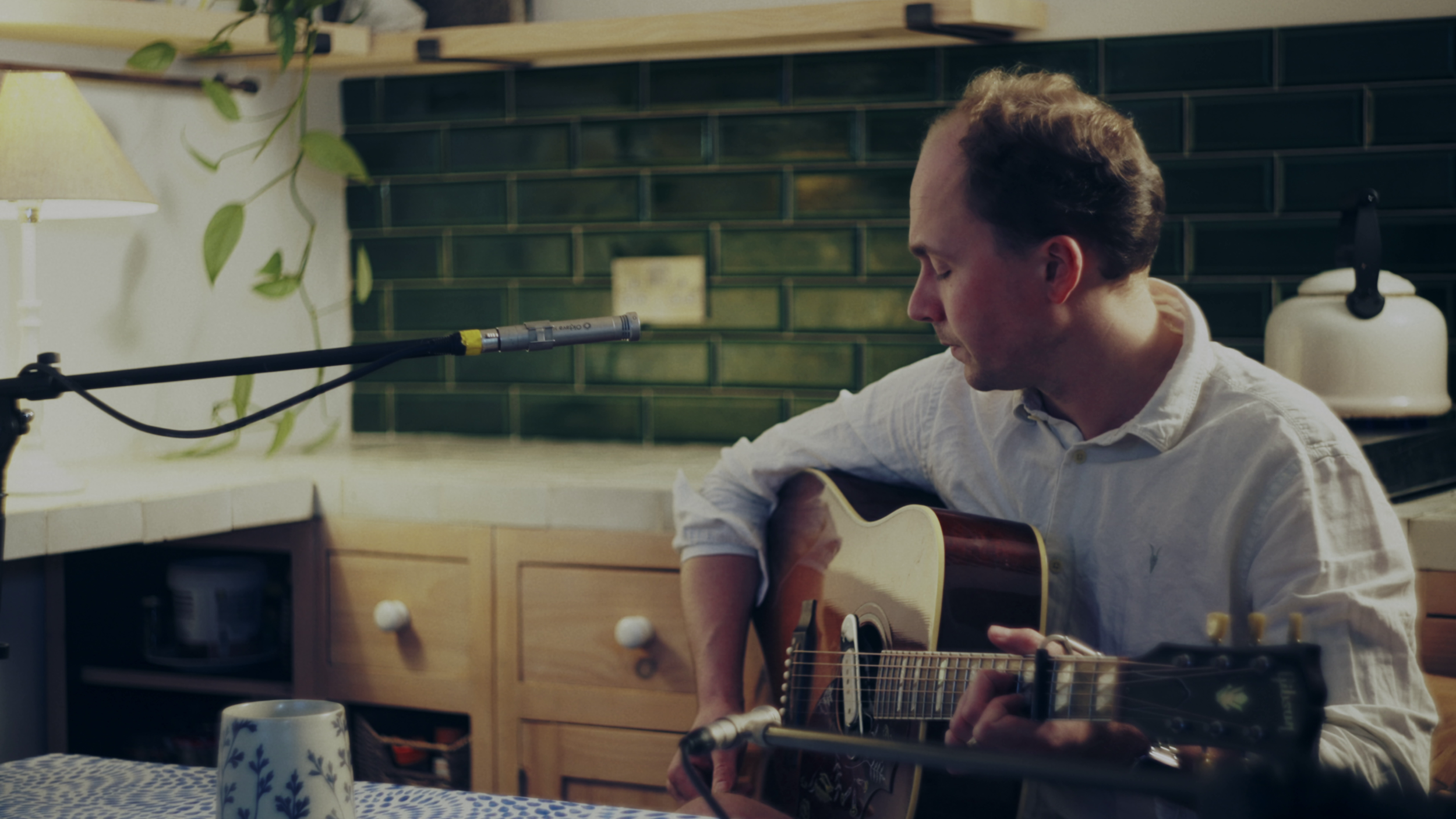 A man with short brown hair playing an acoustic guitar in a cozy kitchen with a dark tiled backsplash, a wooden shelf, a lamp, and a potted plant.