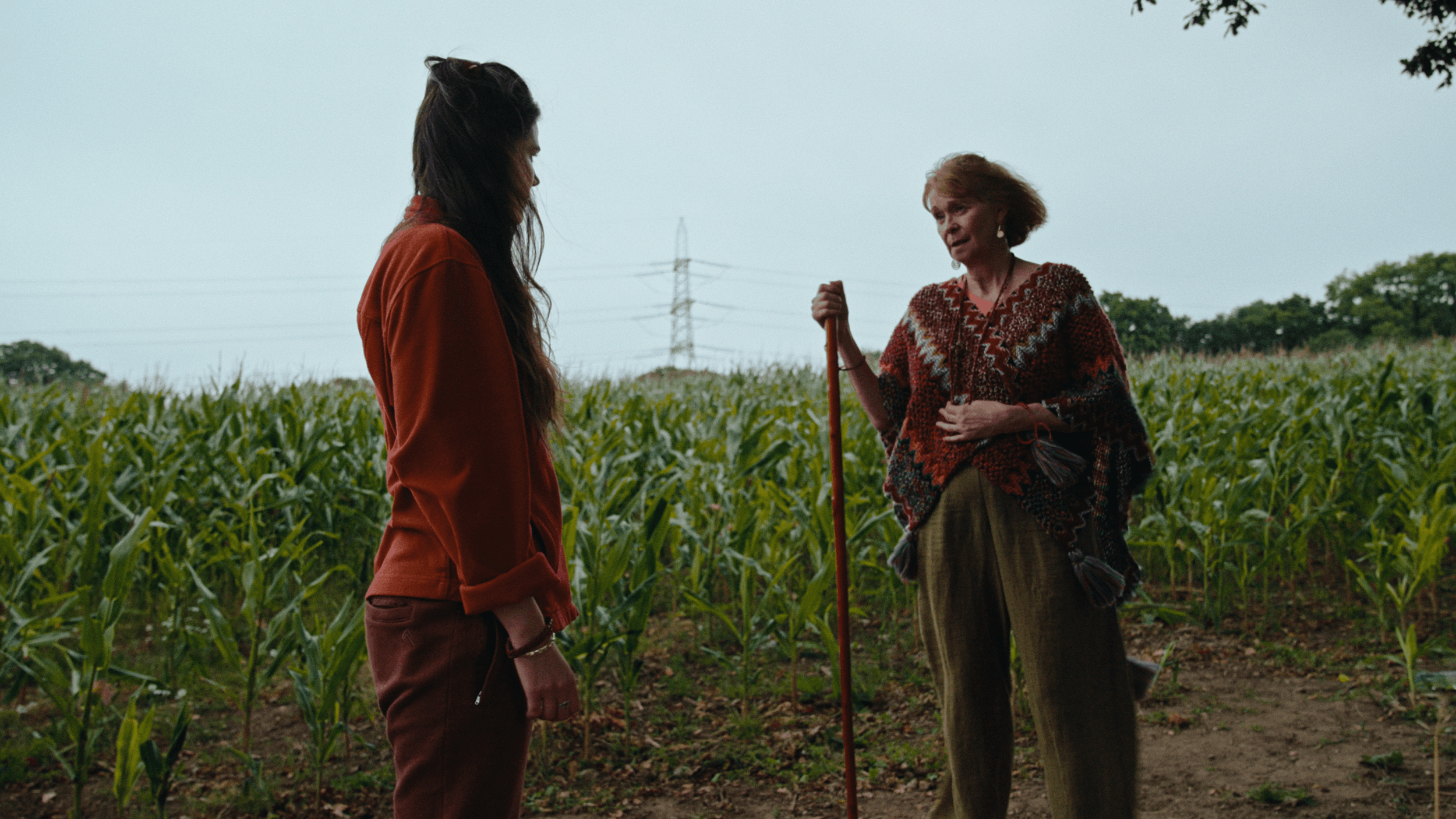 A young woman and an older woman stand in a cornfield having a conversation. The young woman has long dark hair and is dressed in a red jacket and maroon pants. The older woman has short, curly hair and wears a patterned poncho, holding a walking stick.