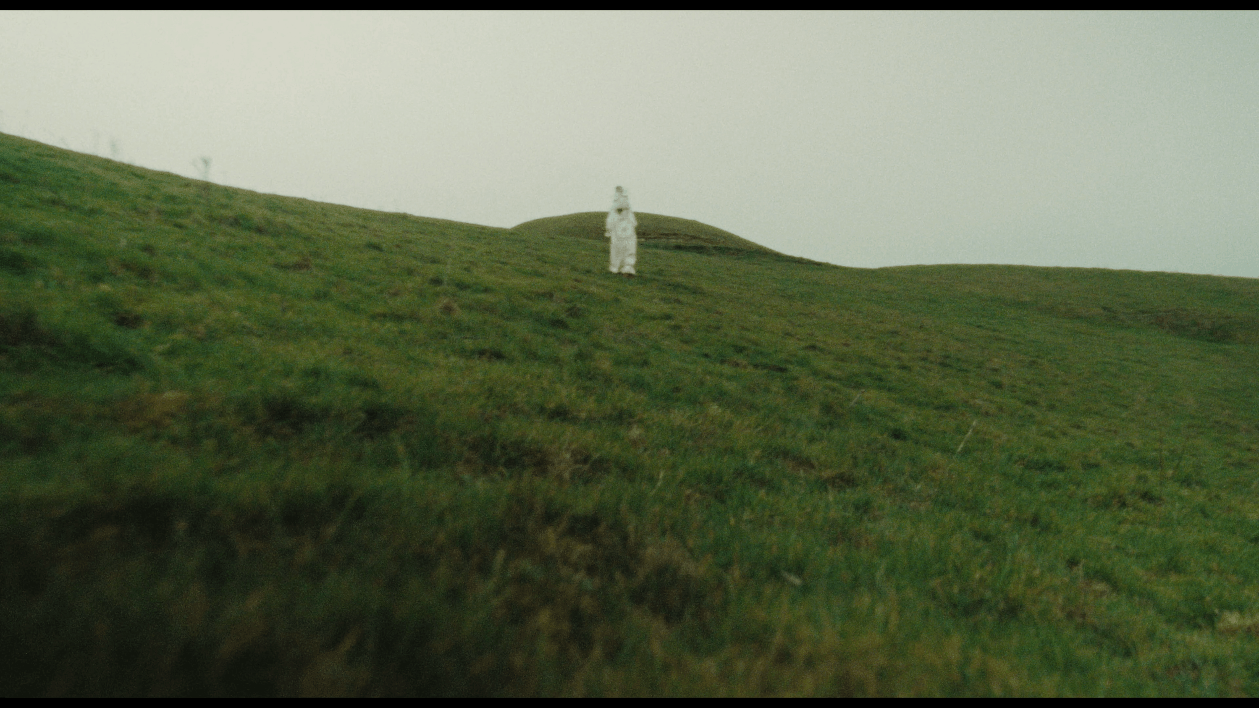 A grassy hill with a cloudy sky, featuring a person wearing white standing at the top.