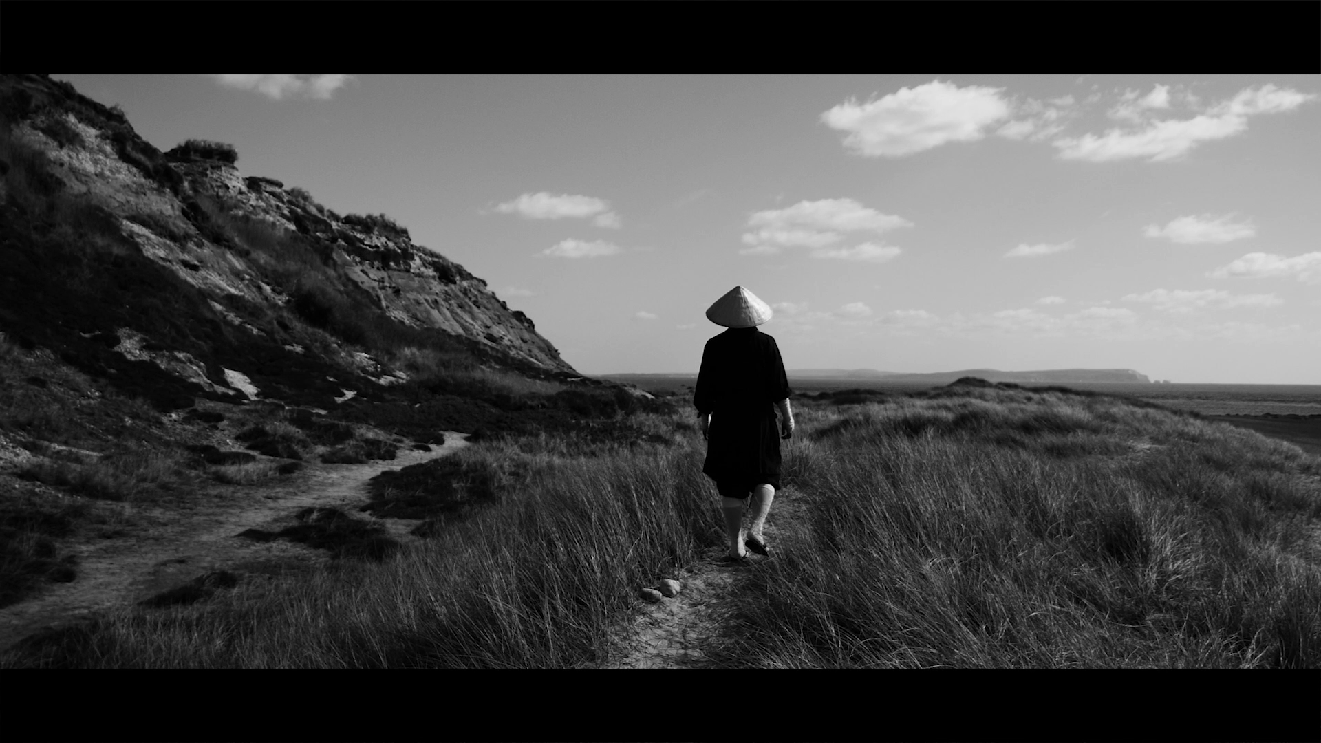 A person wearing a traditional conical hat walking along a dirt path through tall grass, with mountains and clouds in the background, in a black-and-white landscape photograph